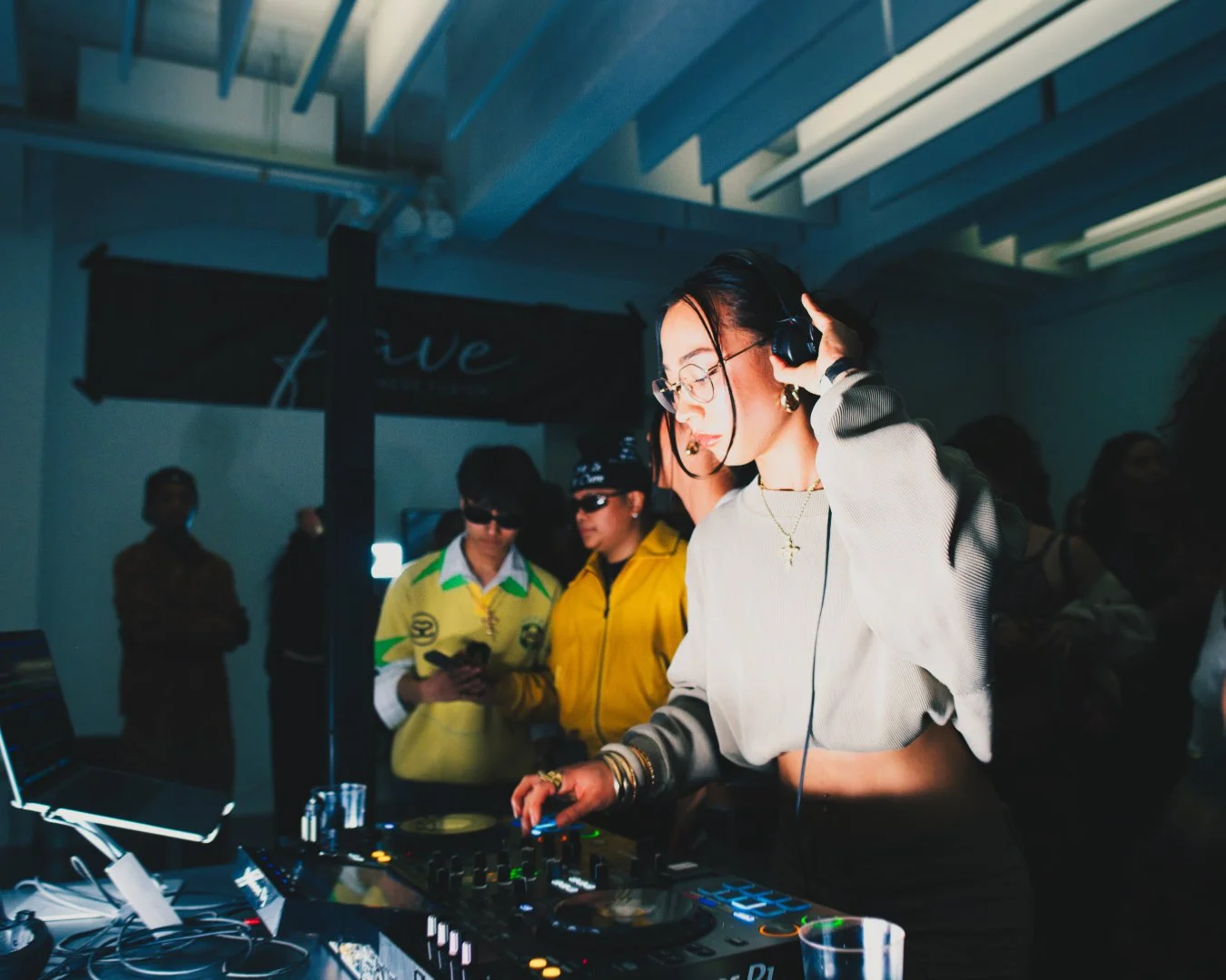 Eine Frau, die als DJ agiert, trägt Kopfhörer und steuert die DJ-Ausrüstung bei einer Party. Im Hintergrund sind Menschen zu sehen, die die Musik genießen