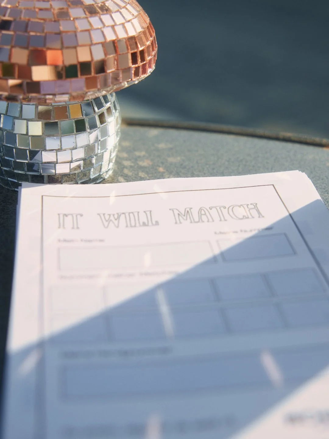 Disco-Kugeln in Gold und Silber neben einem Papier mit dem Text „IT WILL MATCH“ auf einem Tisch im Sonnenlicht