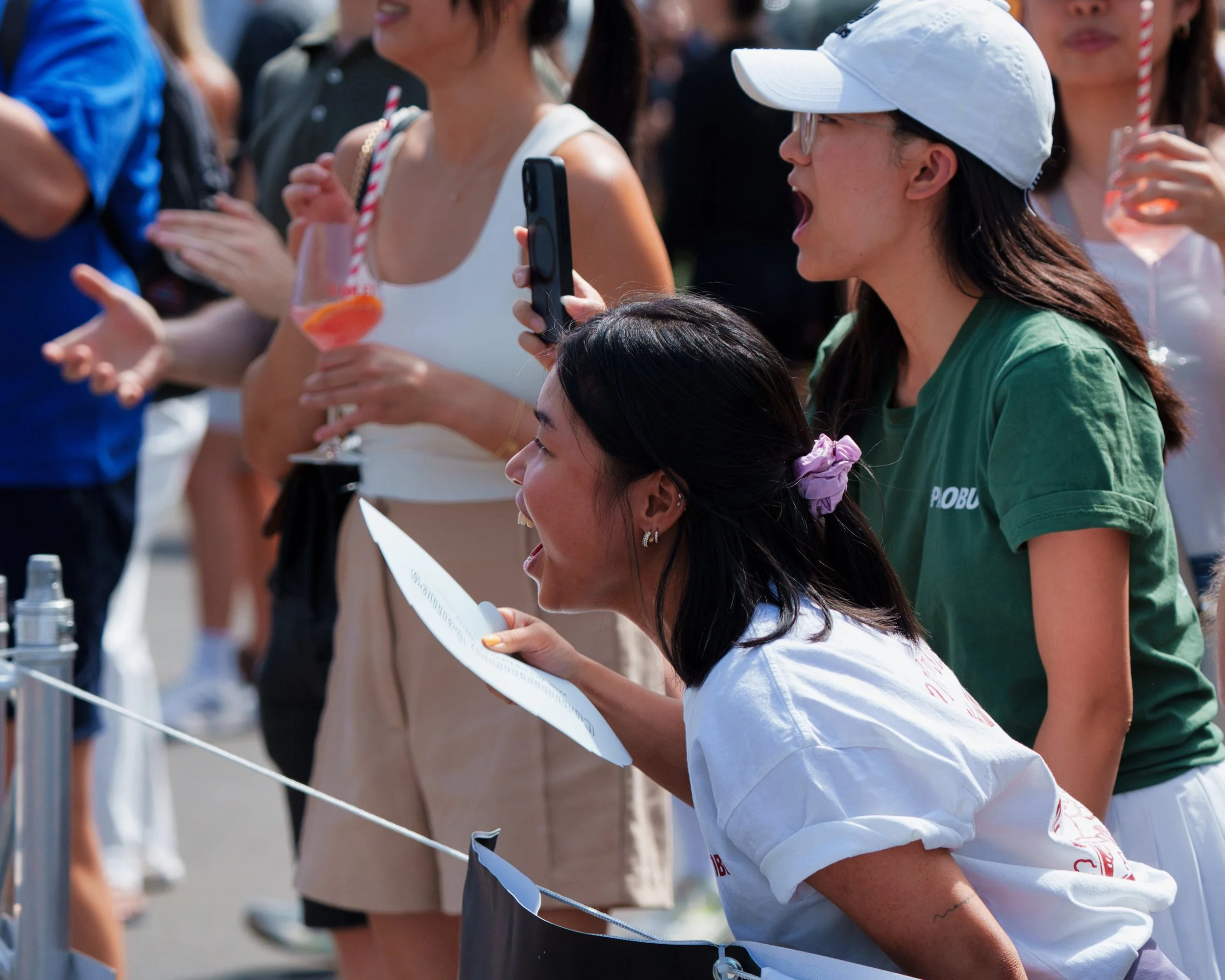 Mehrere Menschen bei einem Event, einige trinken Getränke, eine Person trägt eine weiße Cap und eine Brille, eine person liest ein Blatt, es ist sonnig und warm