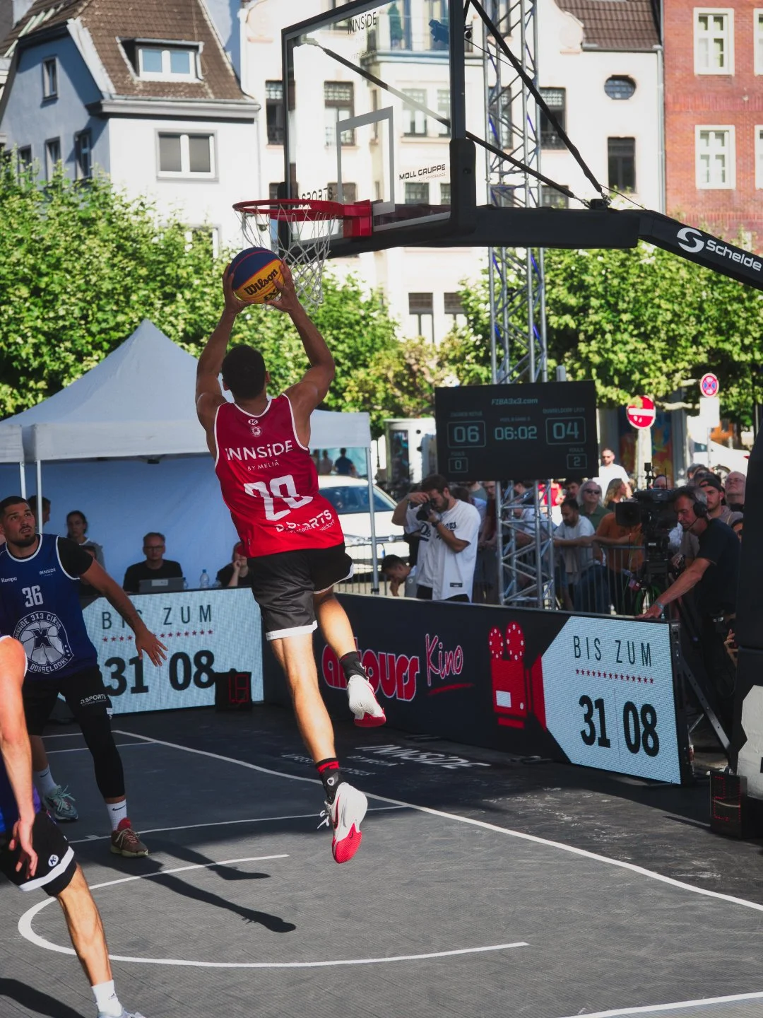 Basketballspieler springt in der Luft, während er den Ball in die Korb wirft bei einem Straßen-Basketballspiel, umgeben von Zuschauern, Kameras und Werbetafeln