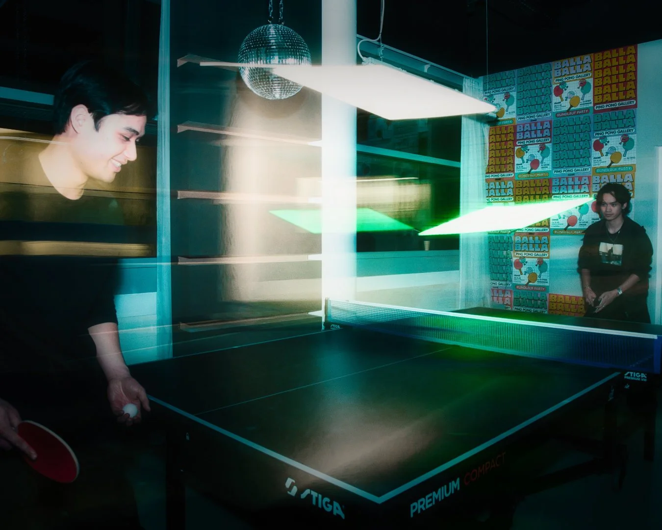 Zwei Menschen spielen Tischtennis in einer Indoor-Spielhalle. Der Hintergrund ist mit bunten Postern zum Thema 'Balla Balla' dekoriert