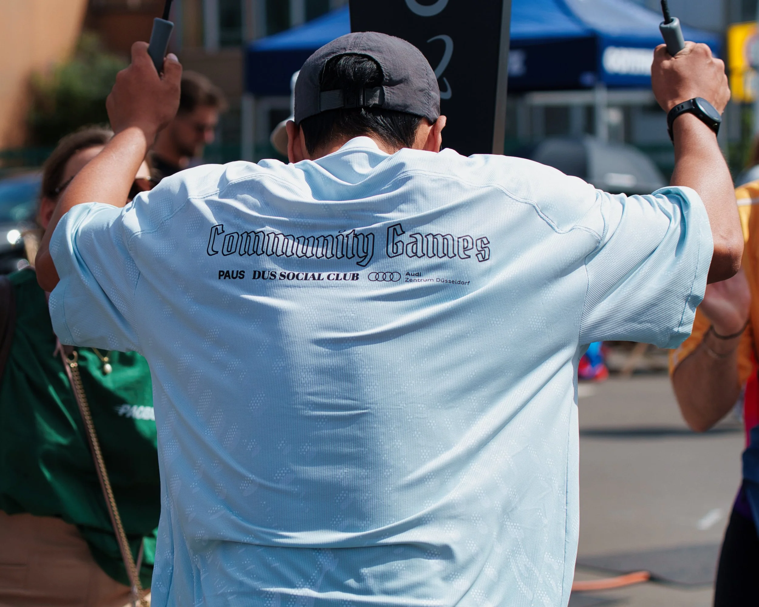 Eine Person trägt ein weißes T-Shirt mit der Aufschrift 'Community Games' und steht in einer Gruppe bei einem gesellschaftlichen Ereignis im Freien