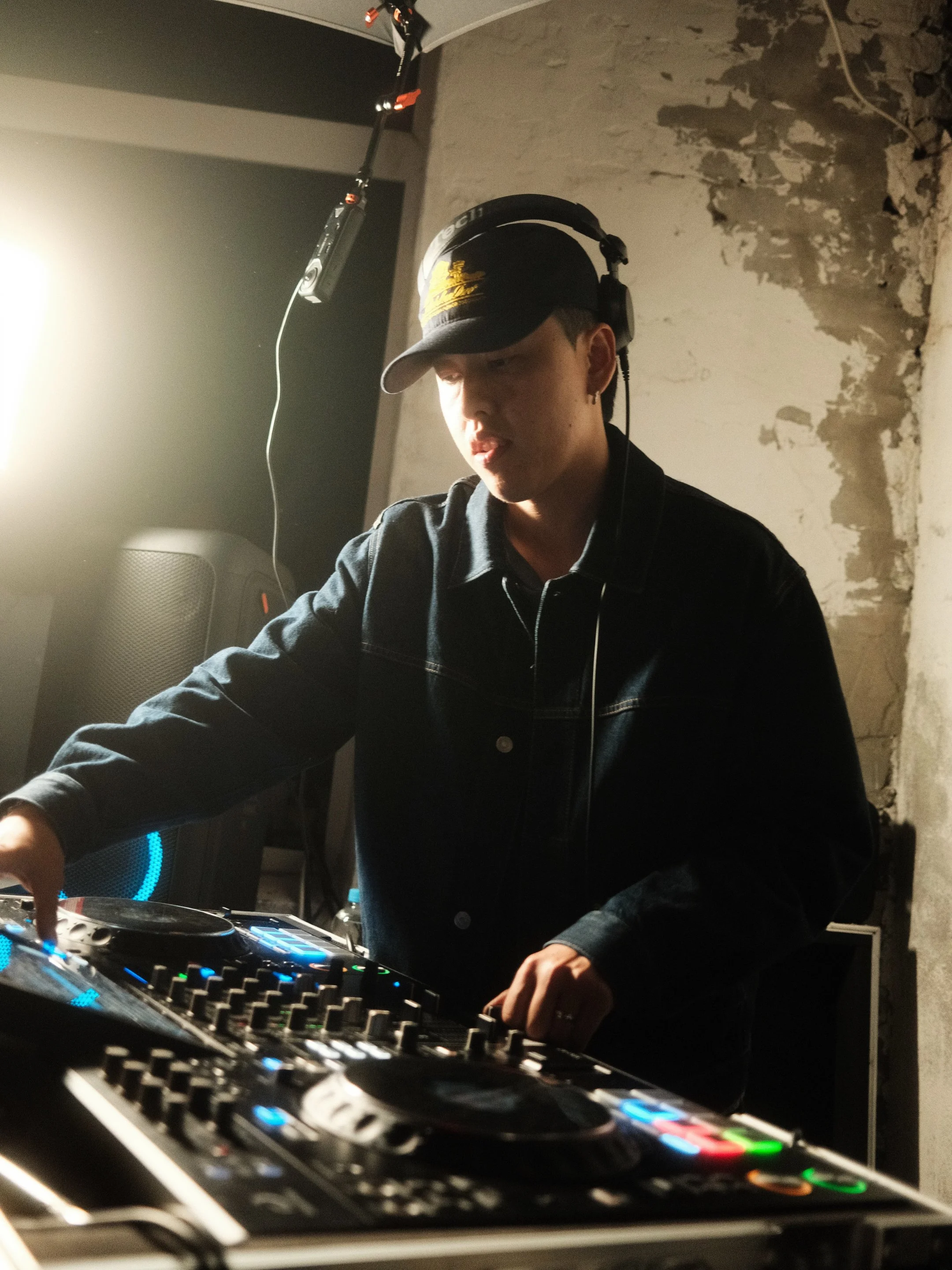Ein junger männlicher DJ in dunkler Kleidung trägt einen schwarzen Baseball-Cap und Kopfhörer, während er an einem DJ-Controller auflegt