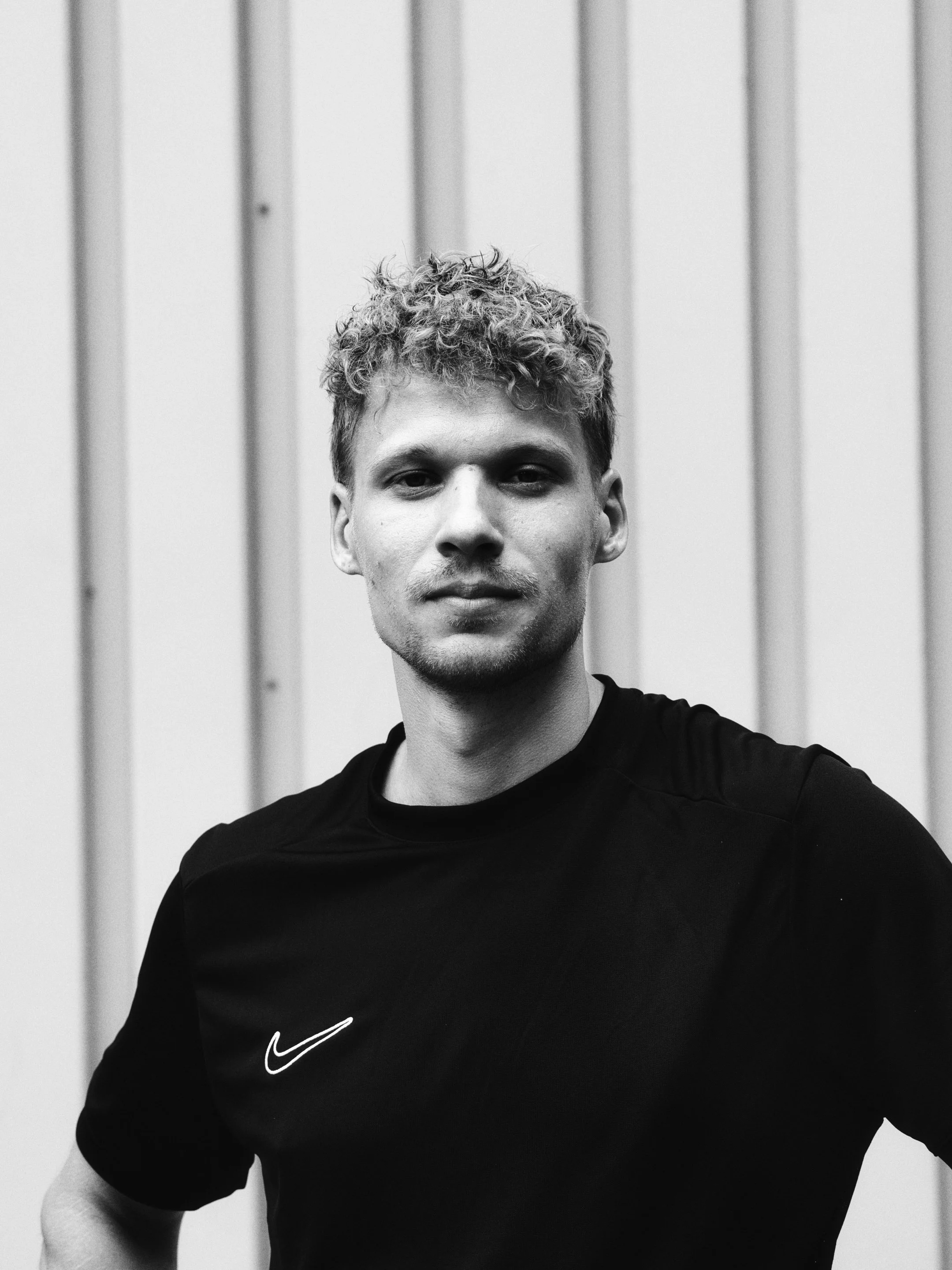 Schwarz-weiß-Fotografie eines jungen Mannes mit lockigem Haar, leichtem Bart und kurzen Haaren, in einem schwarzen Nike-T-Shirt, vor einer vertikal gestreiften Wand
