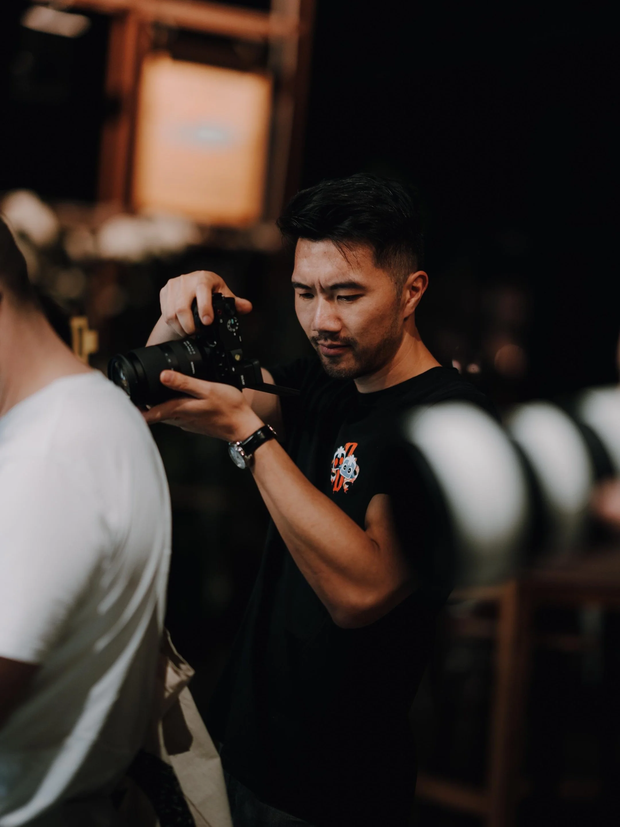 Ein Mann mit schwarzen Haaren und dunklem T-Shirt, das ein lustiges Robotermotiv hat, fotografiert in einem dunklen Raum.