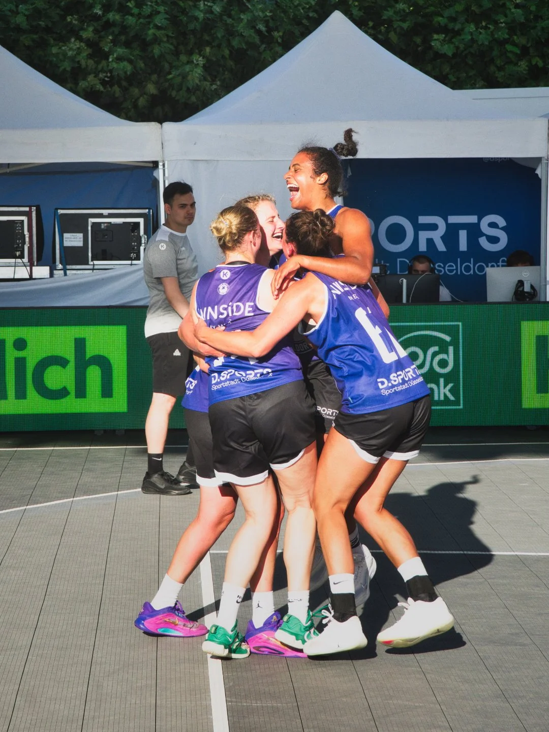 Vier weibliche Volleyballspielerinnen in blauen Trikots und schwarzen Shorts feiern ein Team-Triumph auf einem Basketballfeld, während im Hintergrund ein Mann mit einem weißen T-Shirt und schwarzer Shorts und zwei Männer an einem Tisch sitzen