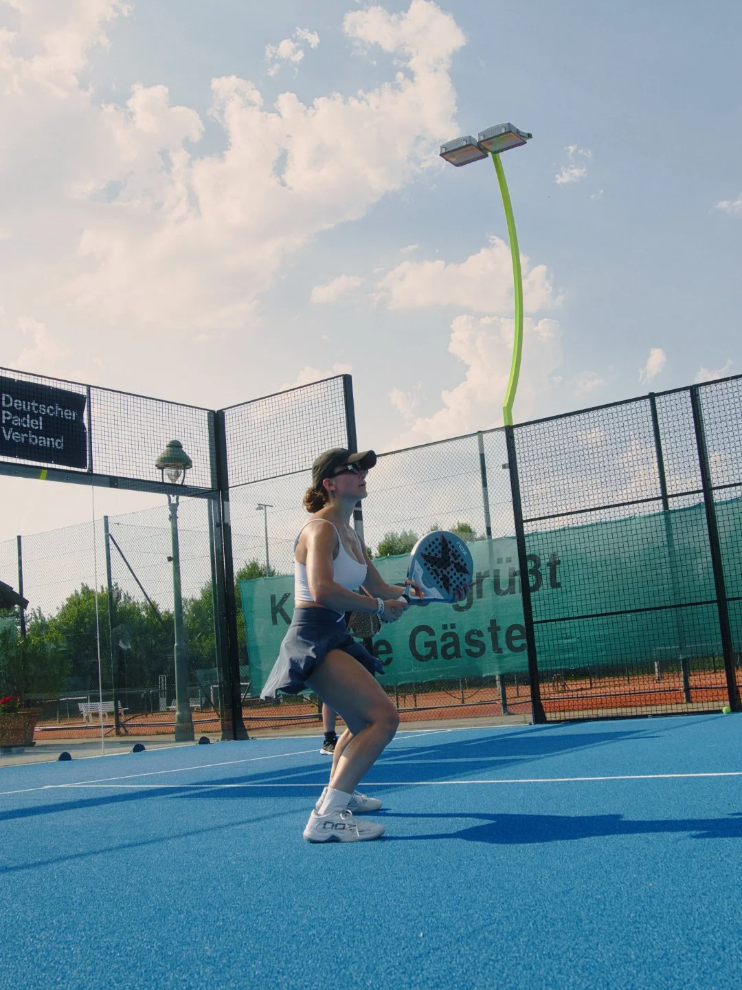 Eine Frau beim Paddel-Tennis auf einem blauen Hartplatz, mit Sonnenbrille und Cap, im Freien bei sonnigem Wetter