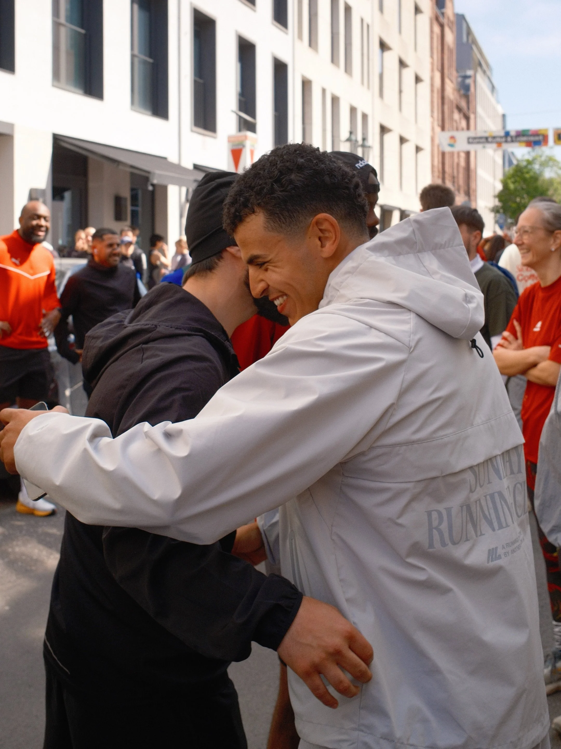 Zwei Männer umarmen sich auf einer Straße mit mehreren Menschen im Hintergrund, die lachen und sprechen, während sie an einem sonnigen Tag draußen sind