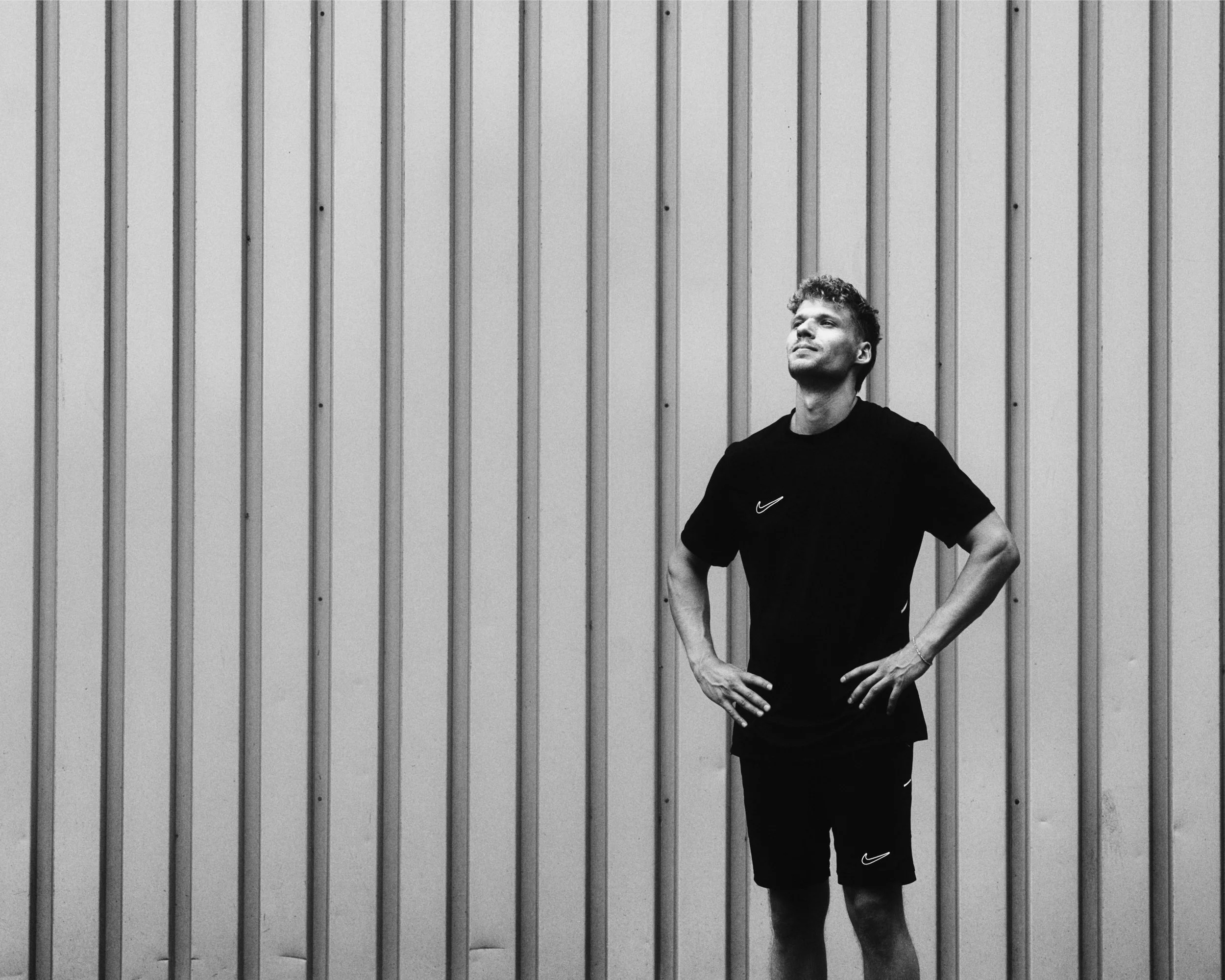 Junge Mann in Sportkleidung steht vor einer vertikalen Metallwand, die nach oben hin verläuft. Er hat die Hände in die Hüften gestützt und blickt nach oben