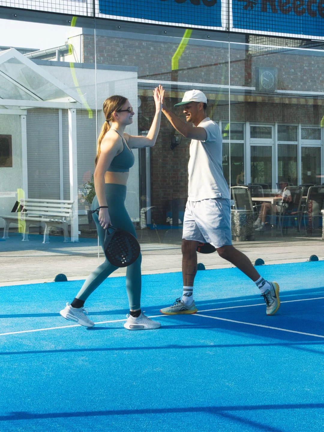 Ein Mann und eine junge Frau beim Padel spielen, die sich auf einem blauen Sportfeld high-fiven, im Hintergrund eine Glaswand und ein Gebäude mit Sitzgelegenheiten