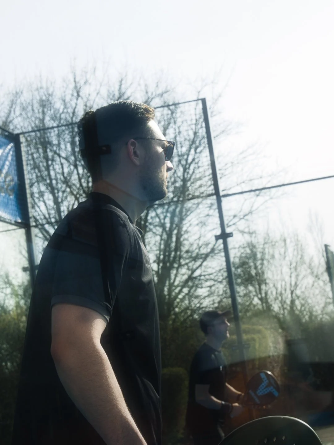 Ein Mann mit dunkler Sonnenbrille und dunklem T-Shirt steht draußen vor einem Tennisplatz, im Hintergrund ein zweiter Mann mit Tennisschläger. eigentlicher Blick nach vorne, Nachmittagssonne, Bäume im Hintergrund