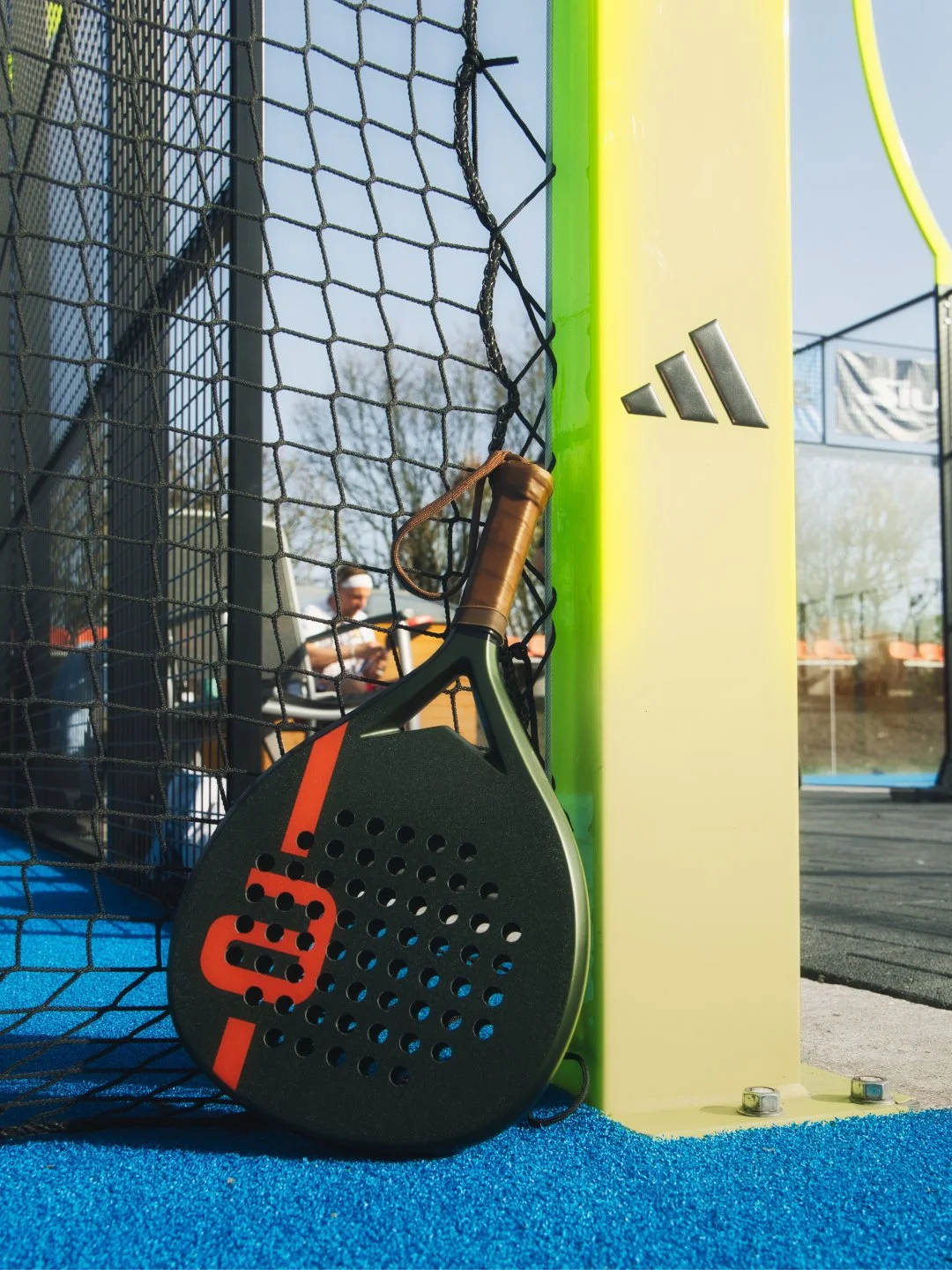 Padel-Schläger vor einem Zaun auf einem Padelplatz, mit einem gelben Pfosten und einer Adidas-Logo im Hintergrund