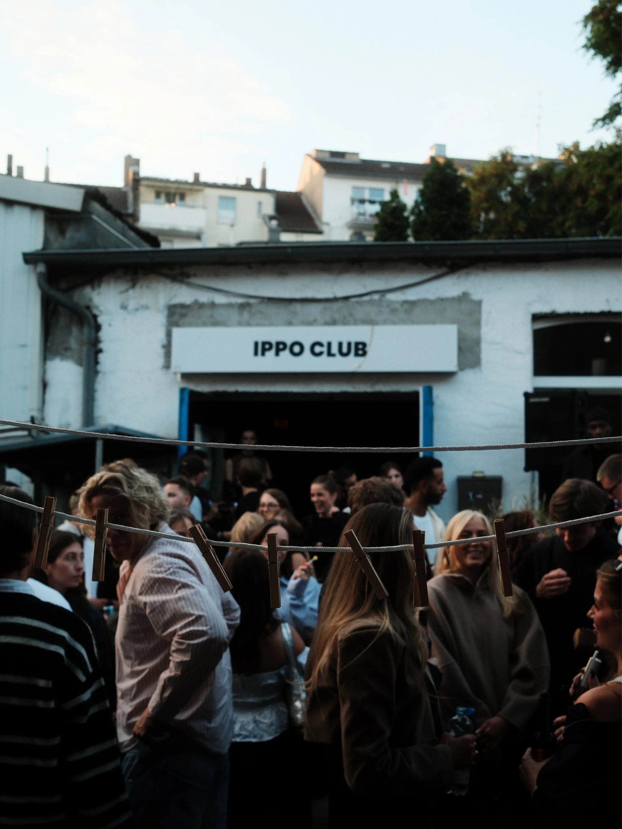 Ein Menschenschwarm vor einem Gebäude mit einem Schild, das 'IPPO CLUB' liest, bei einer Veranstaltung im Freien bei Sonnenuntergang