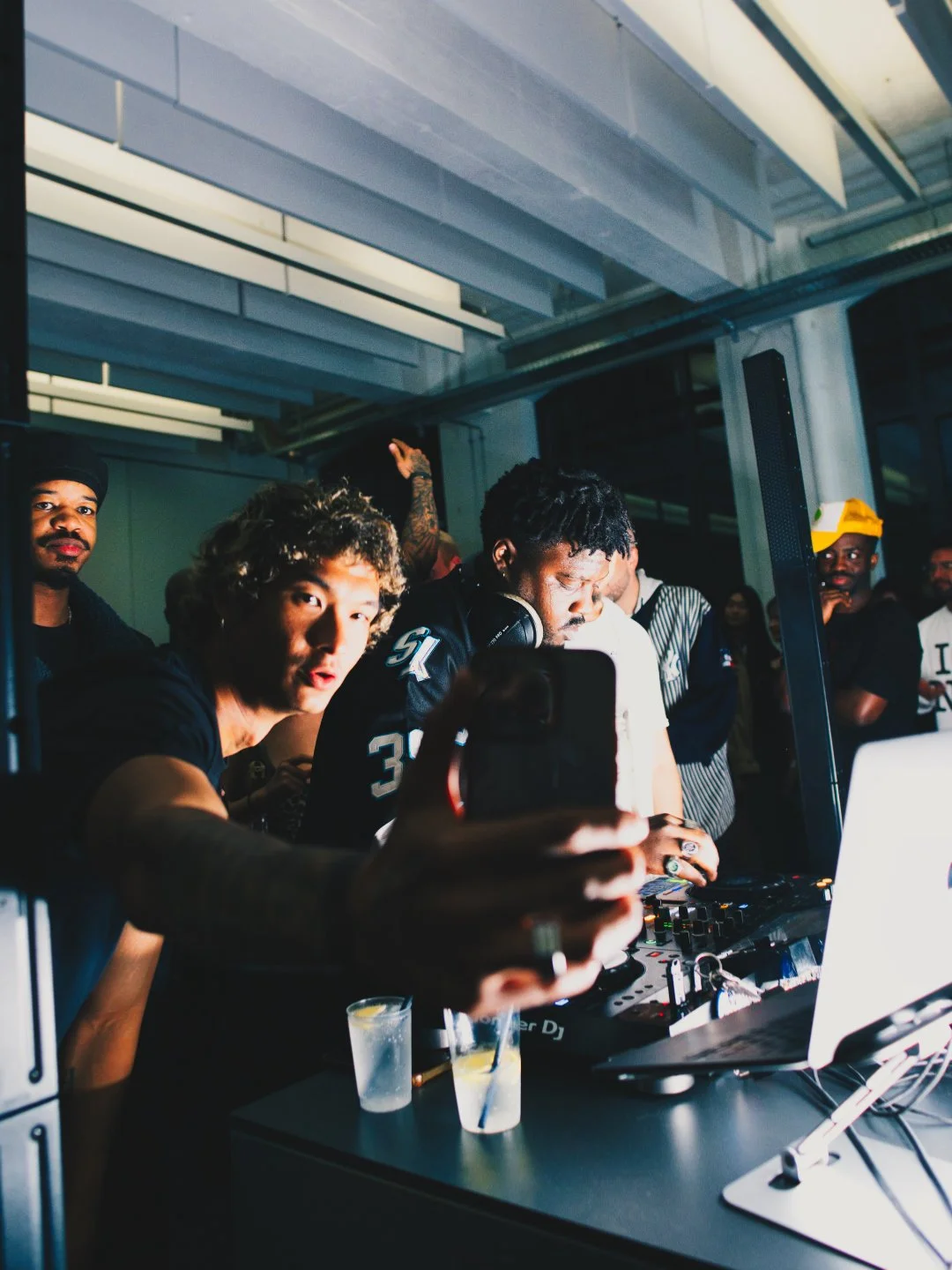 DJ spielt Musik an einem Konzert mit mehreren Zuschauern, Smartphone-Aufnahme, dunkle Innenraum, moderne Architektur, Partystimmung