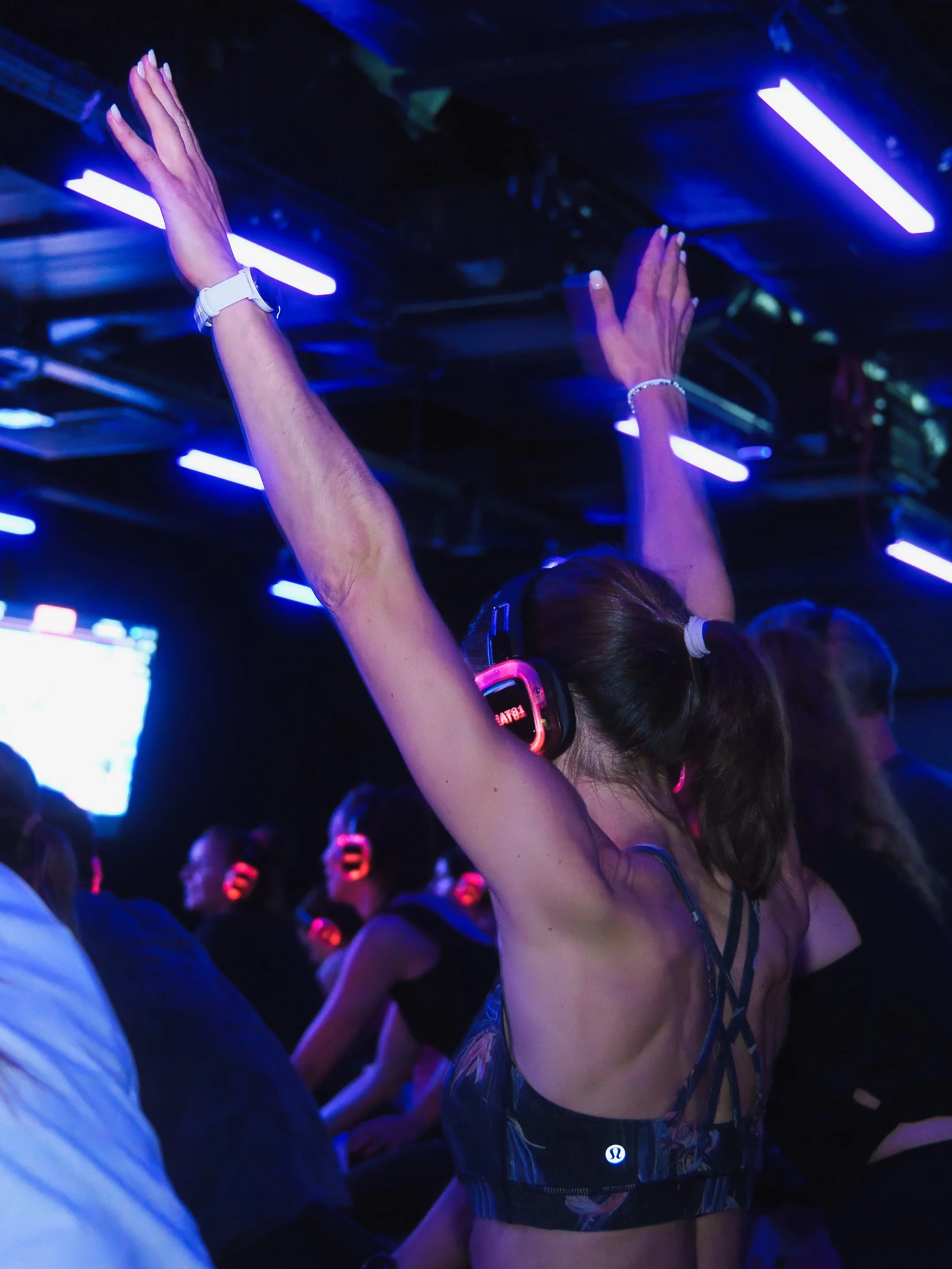 Eine Person im Dunkeln beim Feiern, trägt einen schwarzen Mundschutz, Kopfhörer und ein modisches Top, umgeben von anderen Menschen in einer Party-Atmosphäre mit blauer und violetter Lichtbeleuchtung