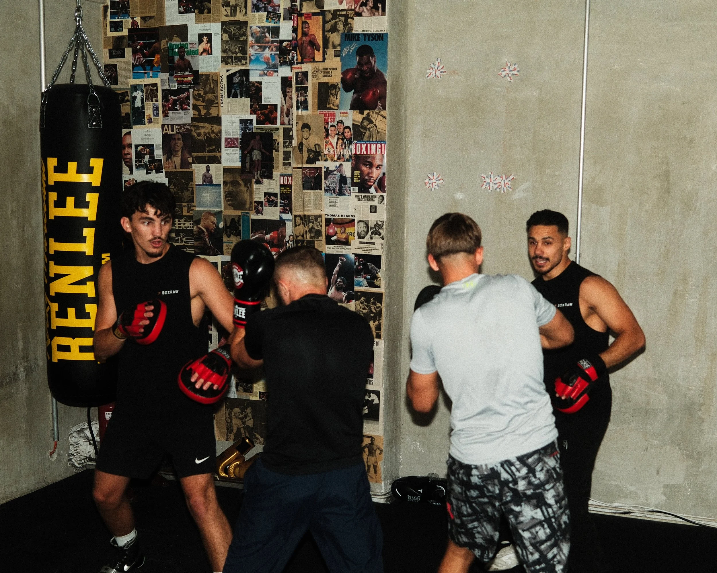 Sieben männliche Boxer in einem Trainingsraum, vier stehen in Trainingskleidung mit Handschuhen, einer hält Boxhandschuhe in der Hand, stehen vor einer Wand mit Boxpostern und Zeitungsausschnitten, es sieht nach einem Boxtraining oder einer Pause aus
