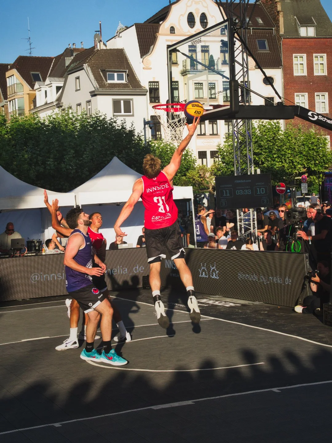 Basketballspieler springt hoch, um Korb vor einer Menschenmenge bei einem Turnier zu werfen, im Hintergrund sind Häuser und ein Publikum