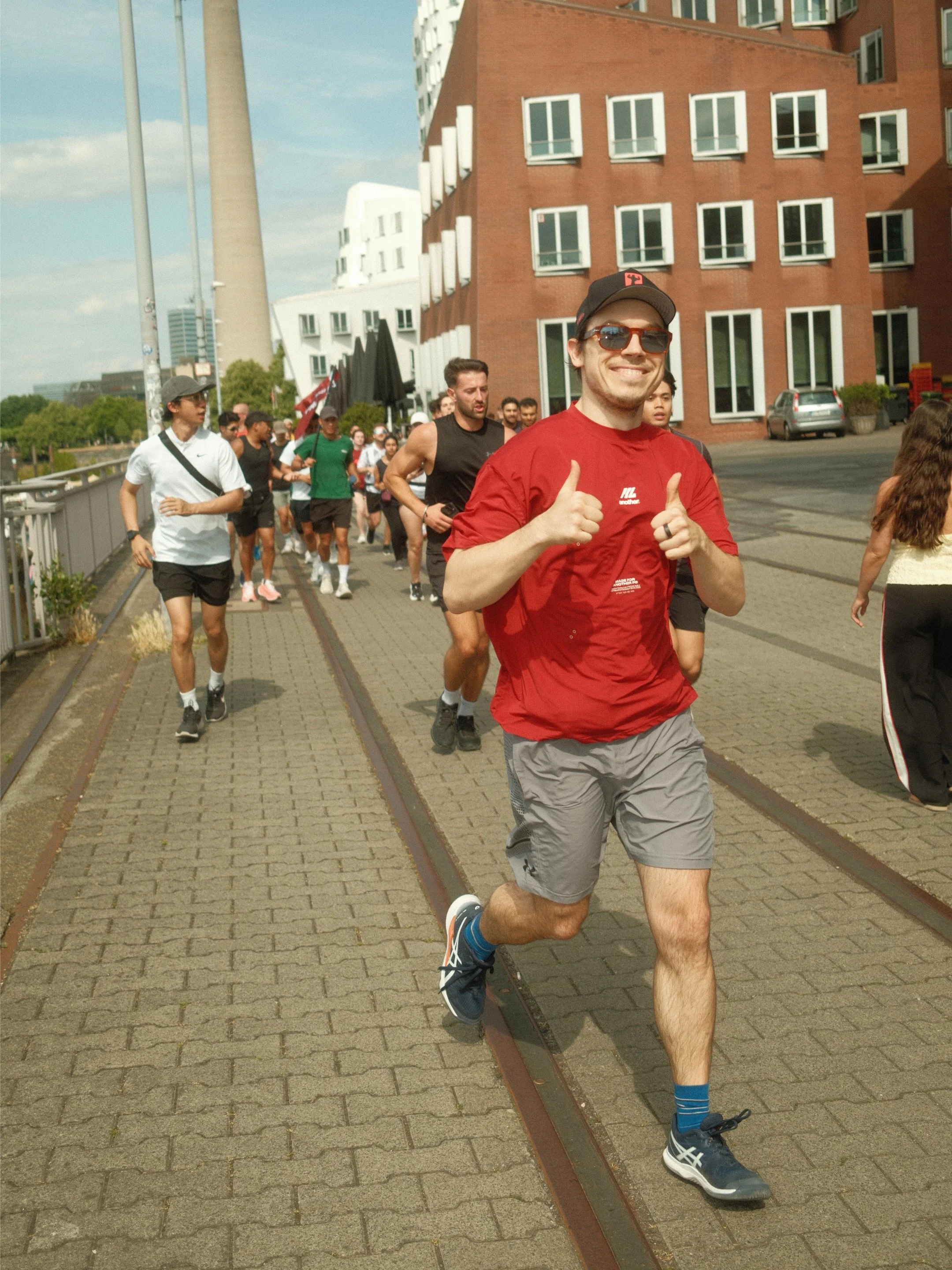 Mann läuft bei einem Straßenlauf, trägt rote T-Shirt, graue Shorts, schwarze Laufschuhe, Sonnenbrille und macht Daumen hoch