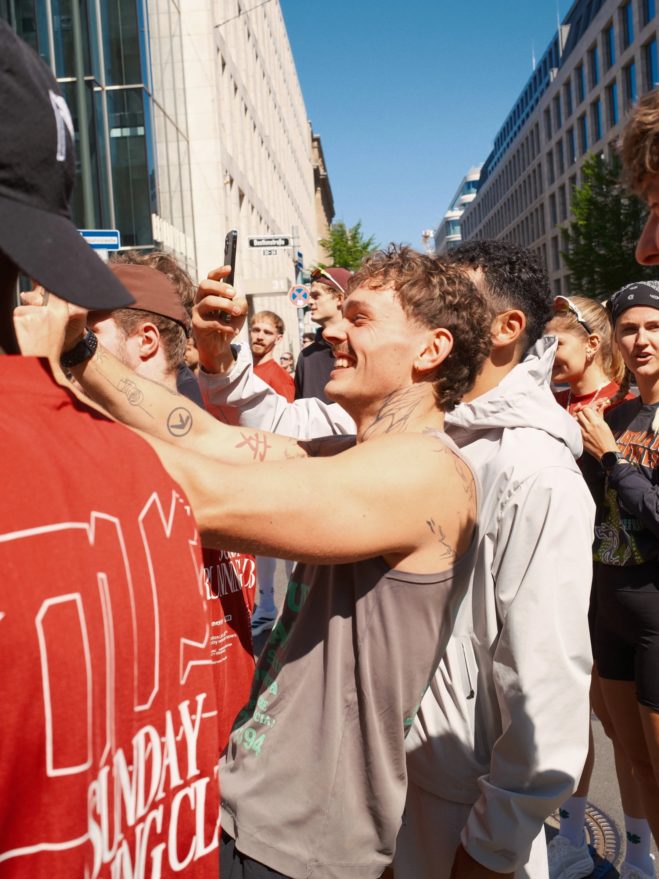 Eine Gruppe von Menschen, die auf einer Stadtstraße stehen und sich gegenseitig begrüßen oder fotografieren. Im Vordergrund ist ein Mann mit Tattoos auf den Armen, der ein Handy in der Hand hält und lächelt