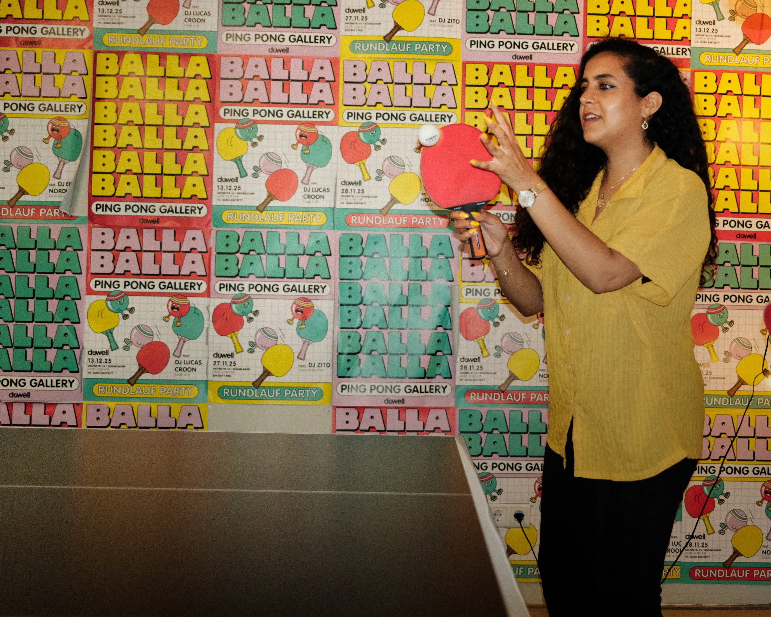 Ein Mann beim Tischtennisspielen vor einem Hintergrund voller bunter Poster mit dem Text 'Balla Balla', 'Ping Pong Gallery' und weiteren Angaben zu Veranstaltungen