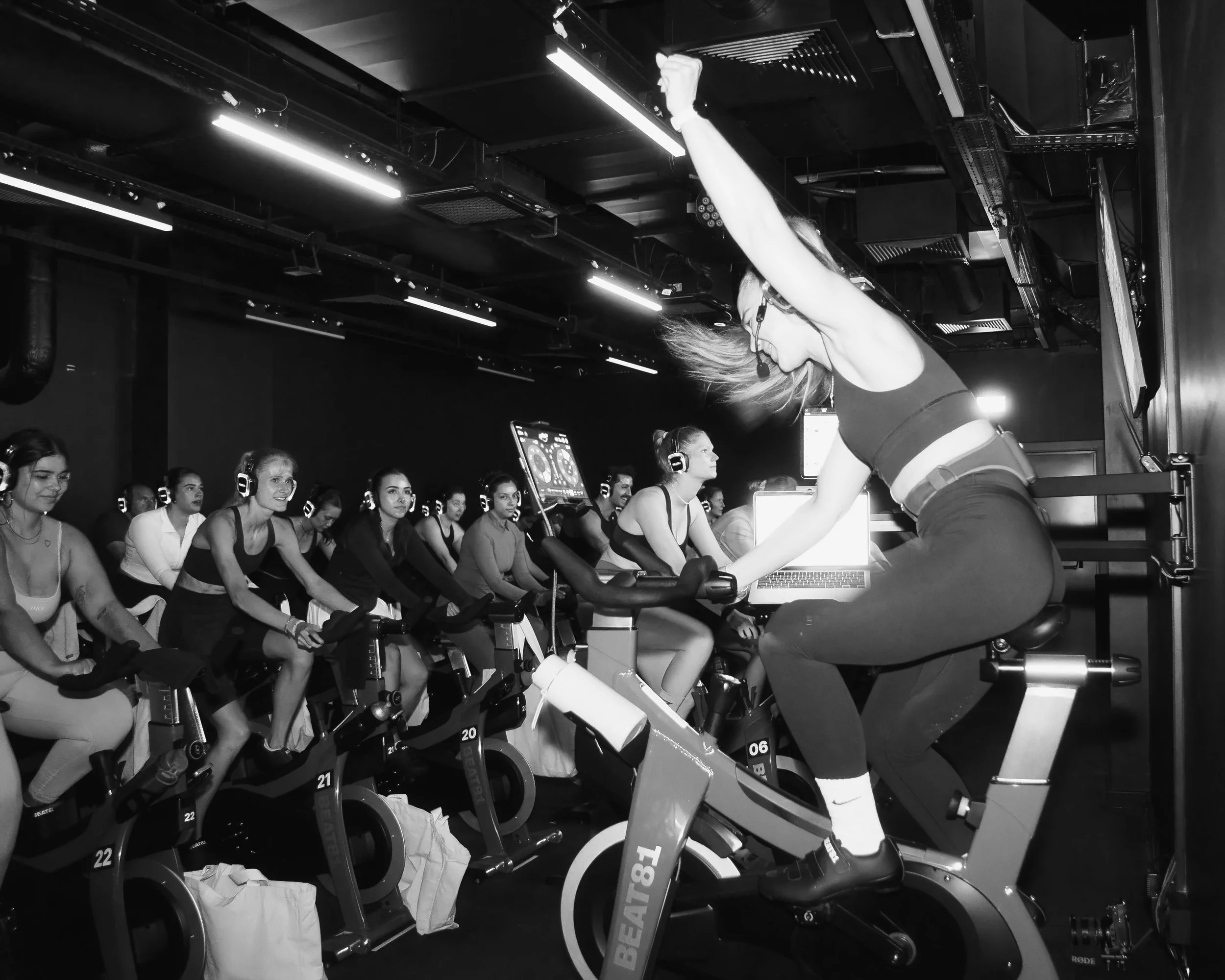 Eine Gruppe von Frauen in einem Fitnessstudio, die an stationären Fahrrädern trainieren. Eine Frau steht in der Mitte, trägt ein Headset und macht eine Übung. Die Szene ist in Schwarz-Weiß