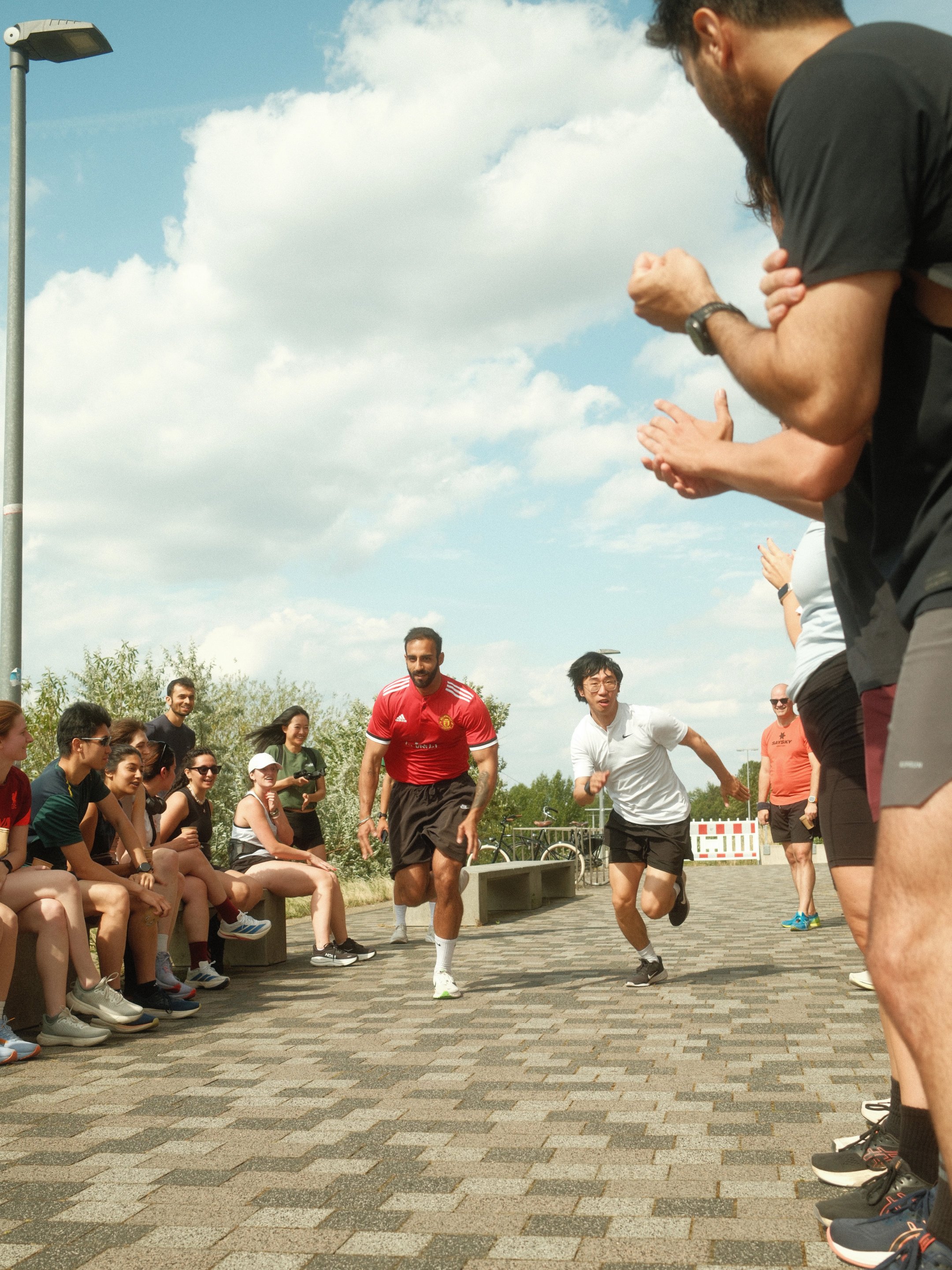 Menschen beim Sprintwettbewerb im Freien, Zuschauer sitzen auf Bänken, einige Fotos machen, der Himmel ist partly cloudy