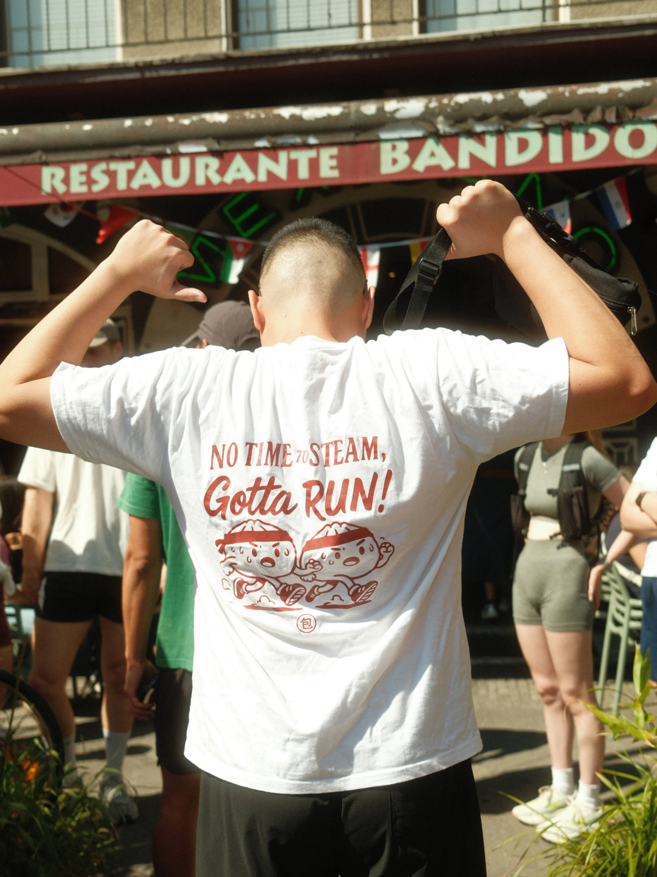 Menschen stehen in einer Menschenmasse vor einem Restaurant mit einem roten und weißen Schild. Die Person im Vordergrund trägt ein weißes T-Shirt mit dem Text 'No Time to Steam, Gotta Run!'.