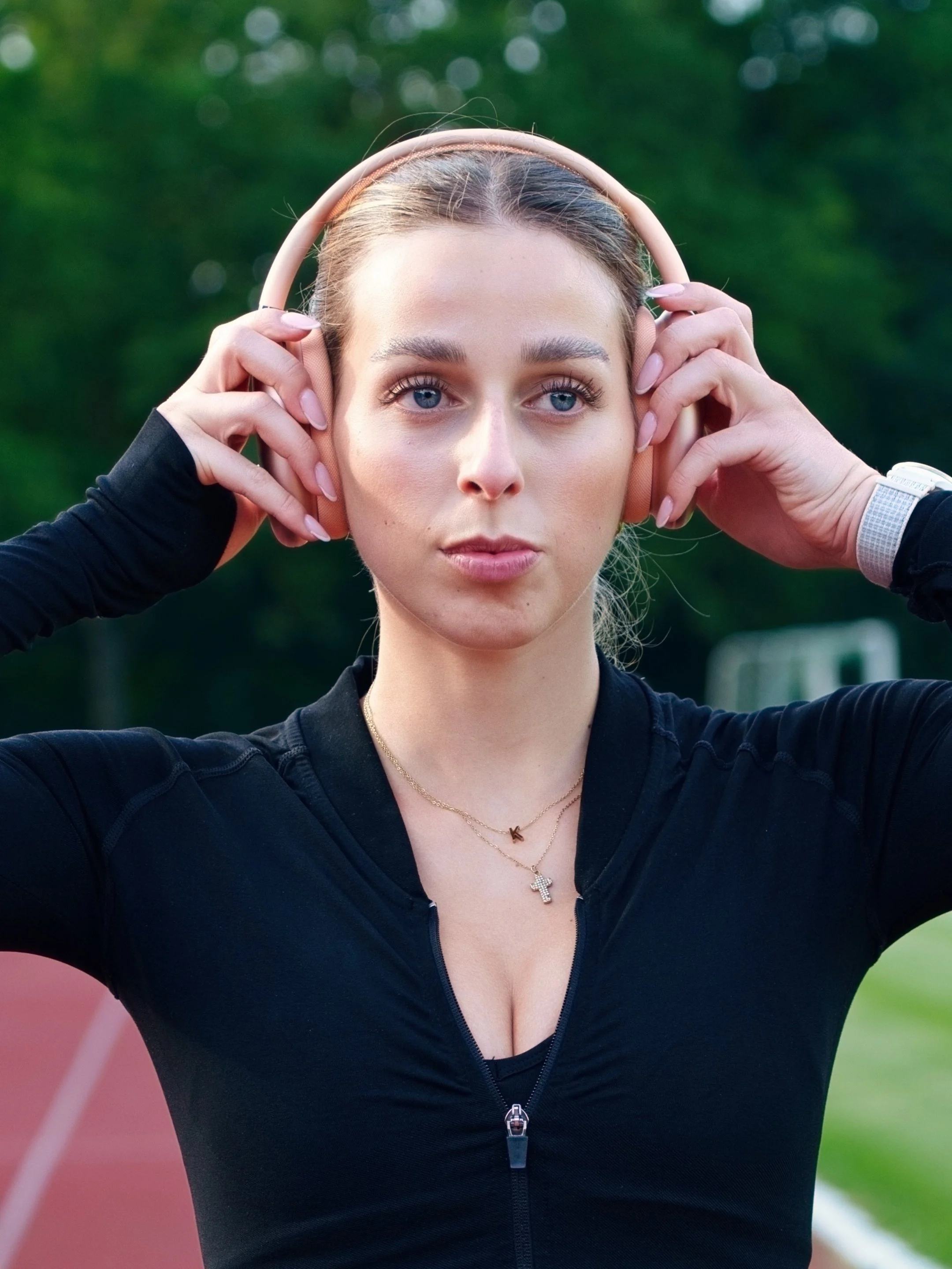 Eine junge Frau mit blauen Augen, blondem Haar, trägt ein schwarz-zipfile Sportoutfit und Kopfhörer, im Hintergrund eine Laufbahn im Freien