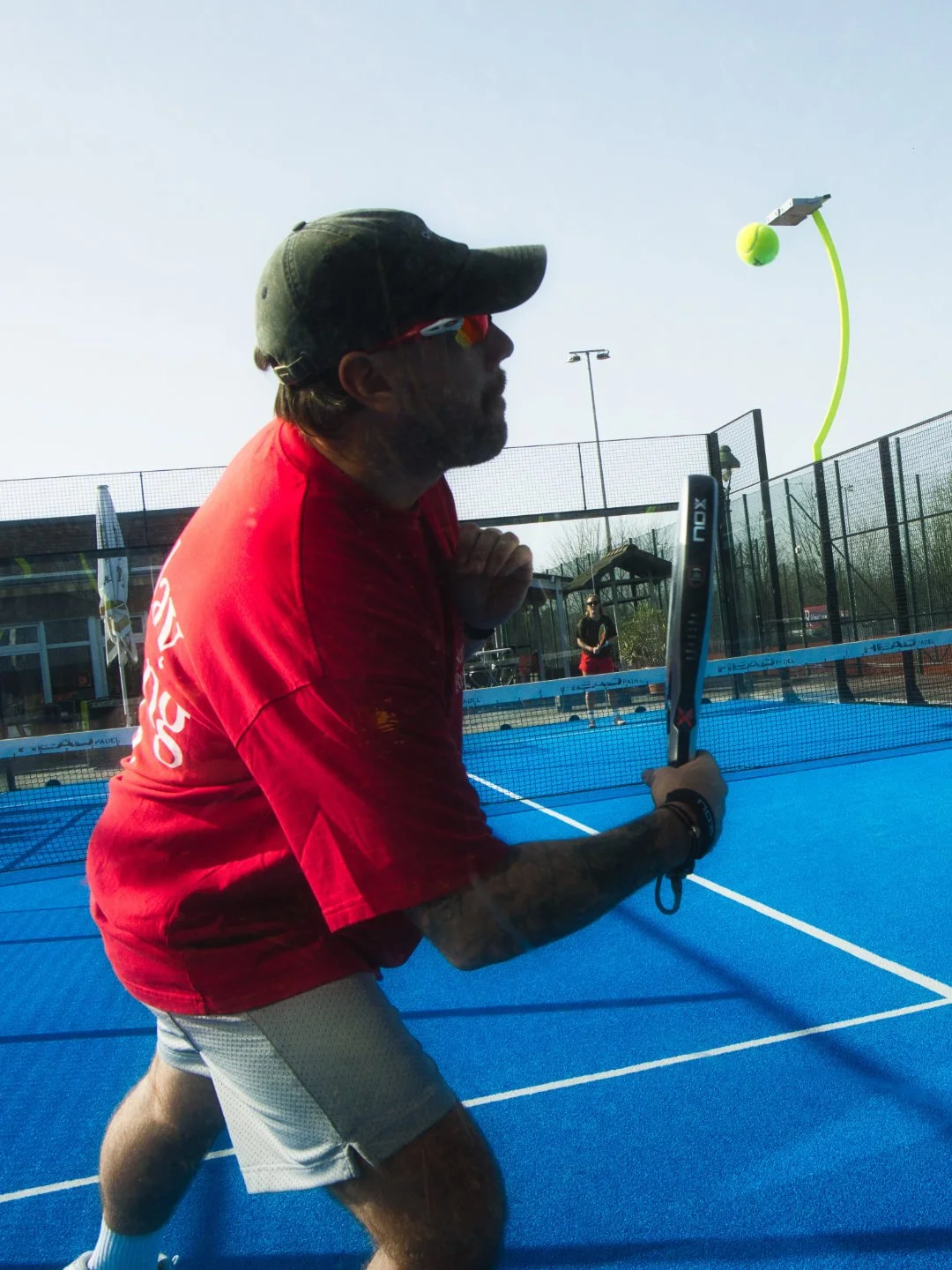 Mann beim Tennis auf dem blauen Tennisplatz, er trägt eine Kappe, Sonnenbrille, rote T-Shirt und graue Shorts, bereitet sich auf einen Schlag vor