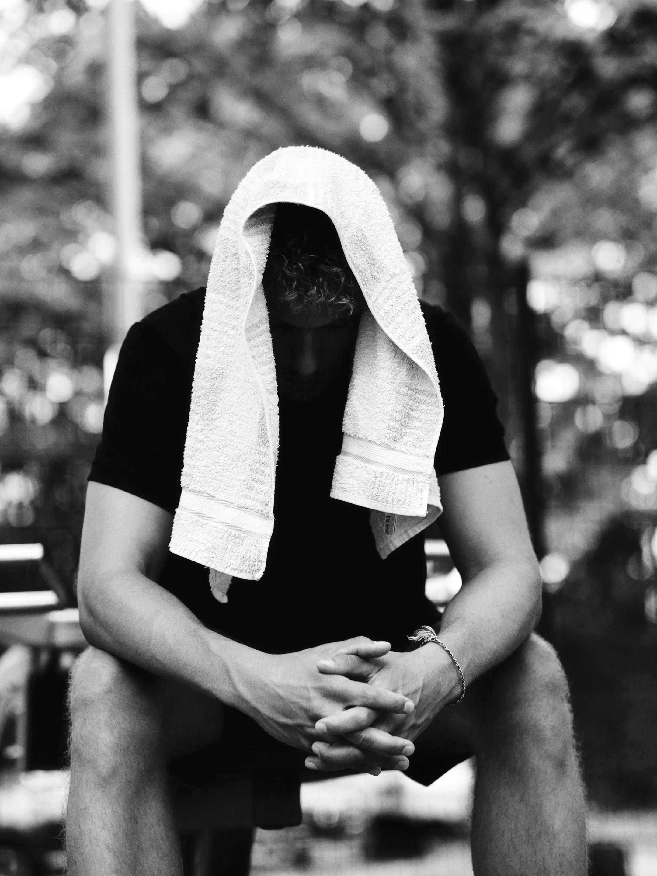 Schwarzweißfoto eines Mannes, der auf einer Bank sitzt, mit einem Handtuch über dem Kopf. Seine Hände sind gefaltet und er trägt eine schwarze T-Shirt, im Hintergrund unscharfe Bäume