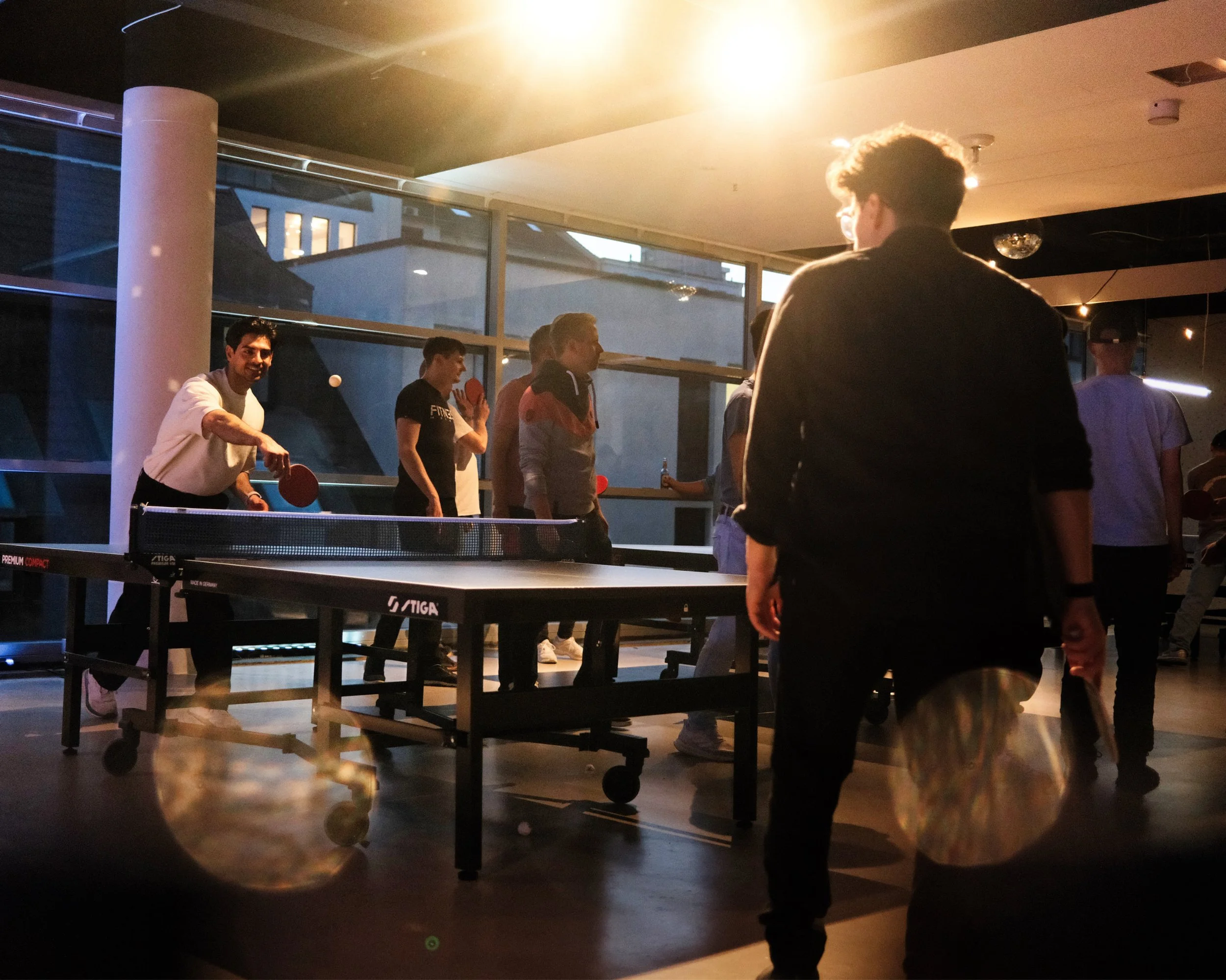 Mehrere Menschen spielen Tischtennis in einem hellen Innenraum bei Abendlicht