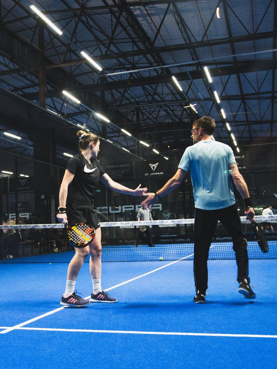 Zwei Menschen auf einem blauen Padelplatz, die sich die Hände schütteln, während sie Tennisschläger halten. Es scheint eine freundliche Geste nach einem Spiel zu sein. Die Halle ist mit künstlichem Licht beleuchtet