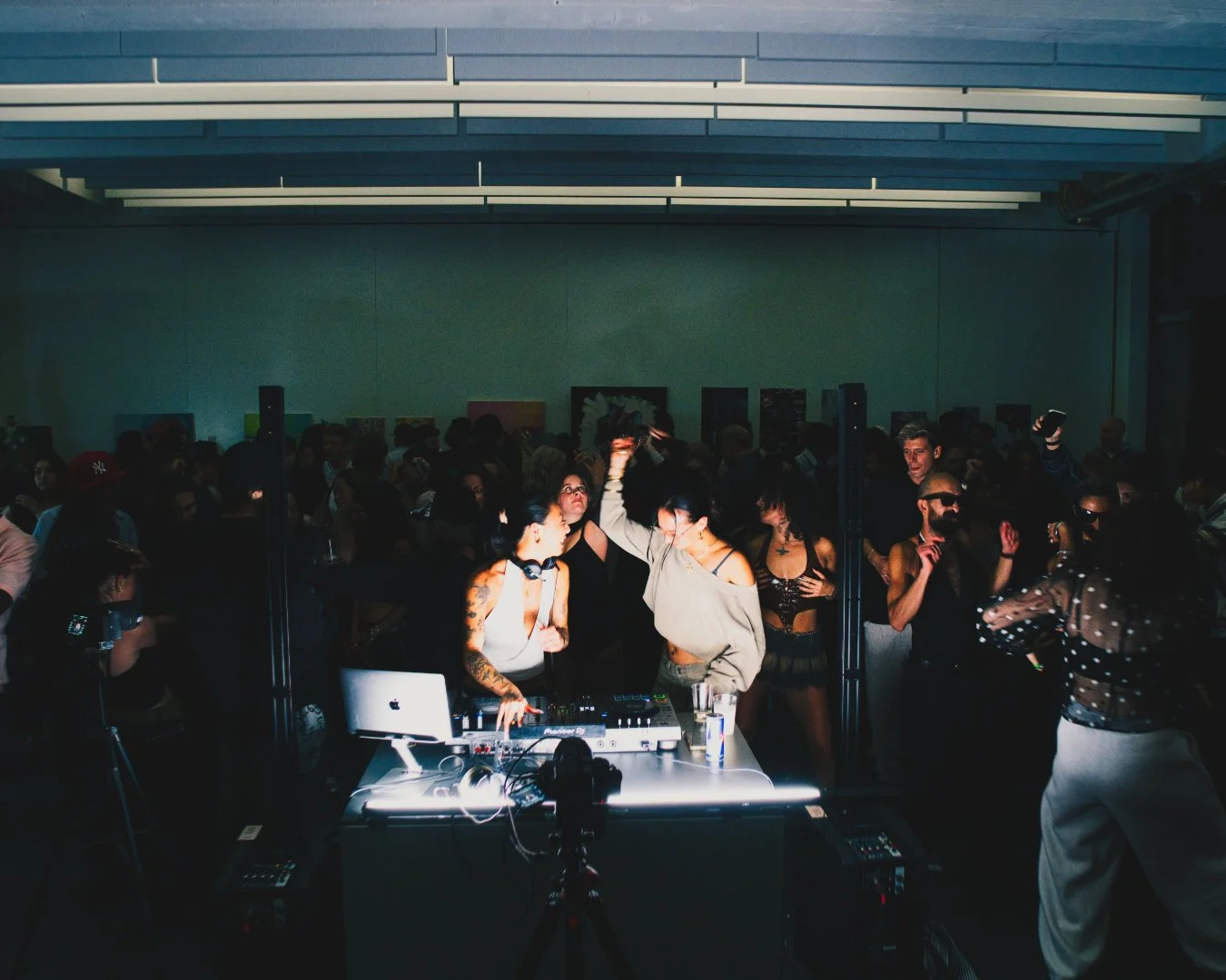 Menschen feiern in einer dunklen, modernen Club-Umgebung mit Disco-Beleuchtung, Tanzen und Musik, einige tragen Sonnenbrillen, ein DJ ist im Vordergrund mit einem DJ-Equipment