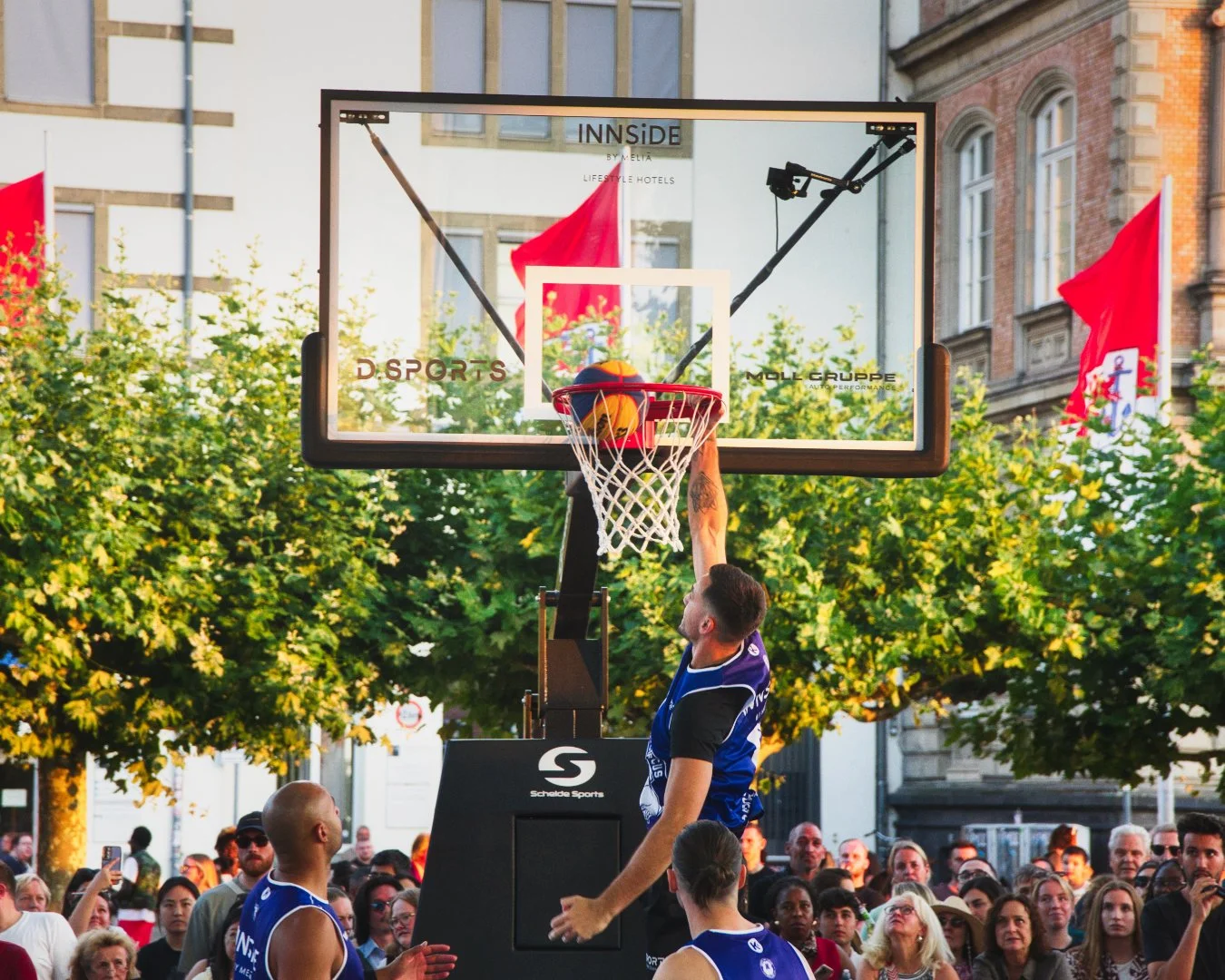 Mann springt beim Basketballspiel, wirft einen Ball in den Korb, mit einem Publikum im Hintergrund, in einer Stadt, bei Sonnenuntergang