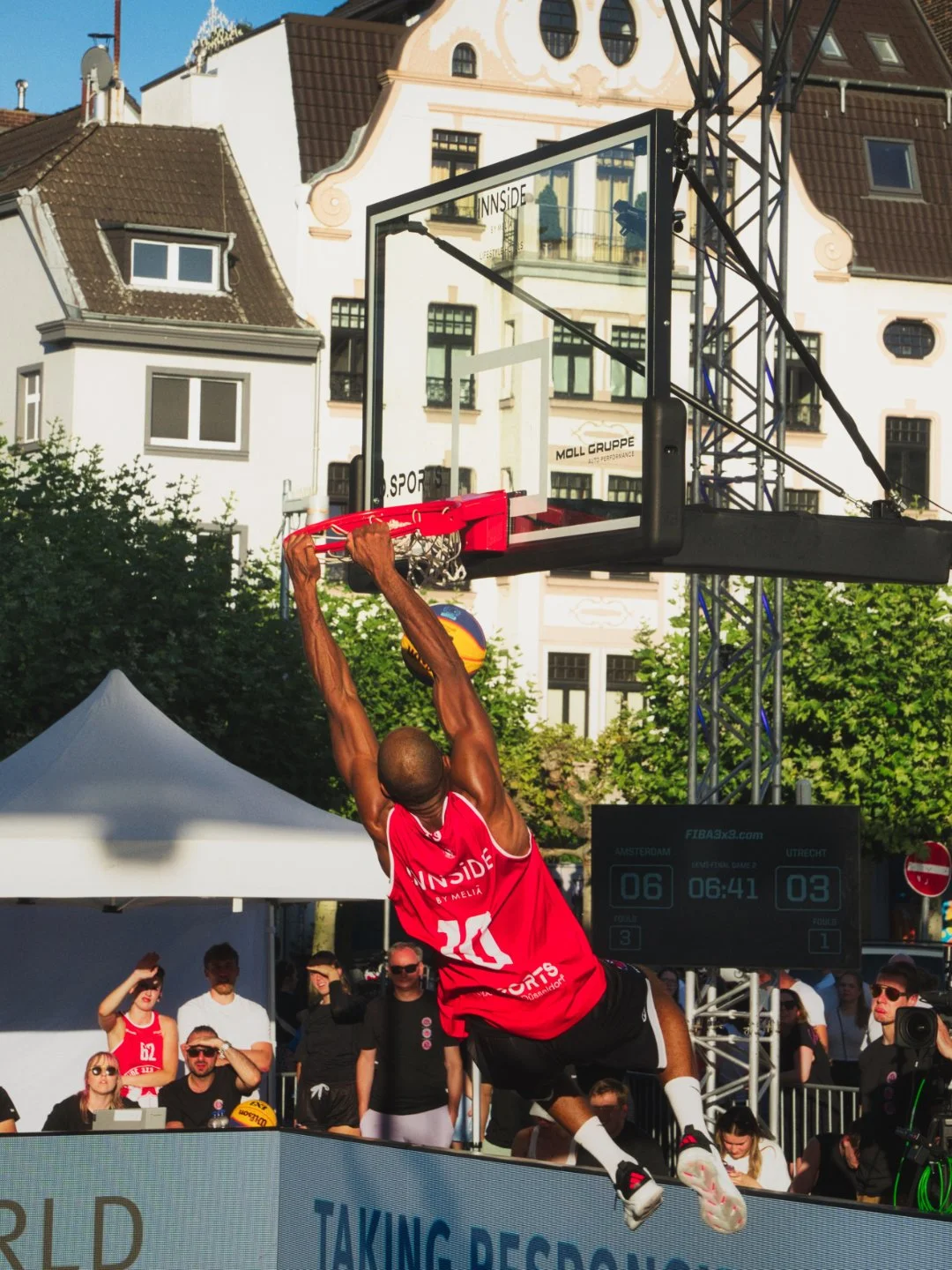 Basketballspieler in roten Trikots springt zum Dunken gegen den Korb bei einem Straßenbasketballturnier, mit Zuschauern im Hintergrund und einem digitalen Spielstand auf dem Bildschirm