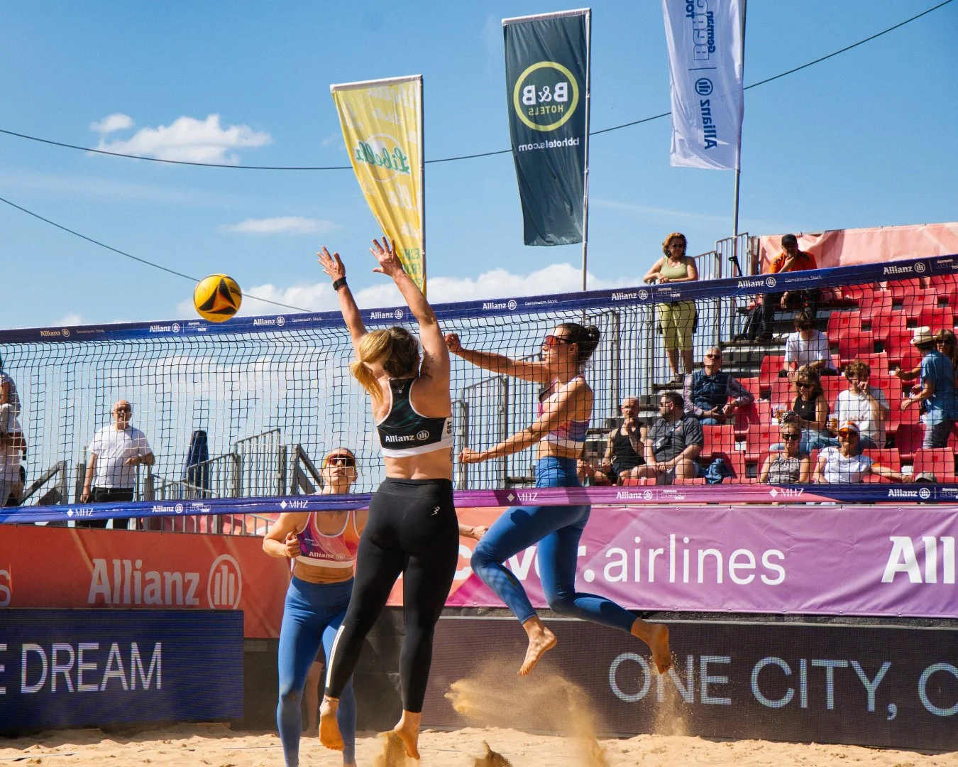 Zwei Frauen beim Beachvolleyball im Wettkampf, mit einem Netz zwischen ihnen, während die anderen zuschauen, auf einer Strandboden. Es ist sonnig und es gibt mehrere Zuschauer und Flaggen im Hintergrund