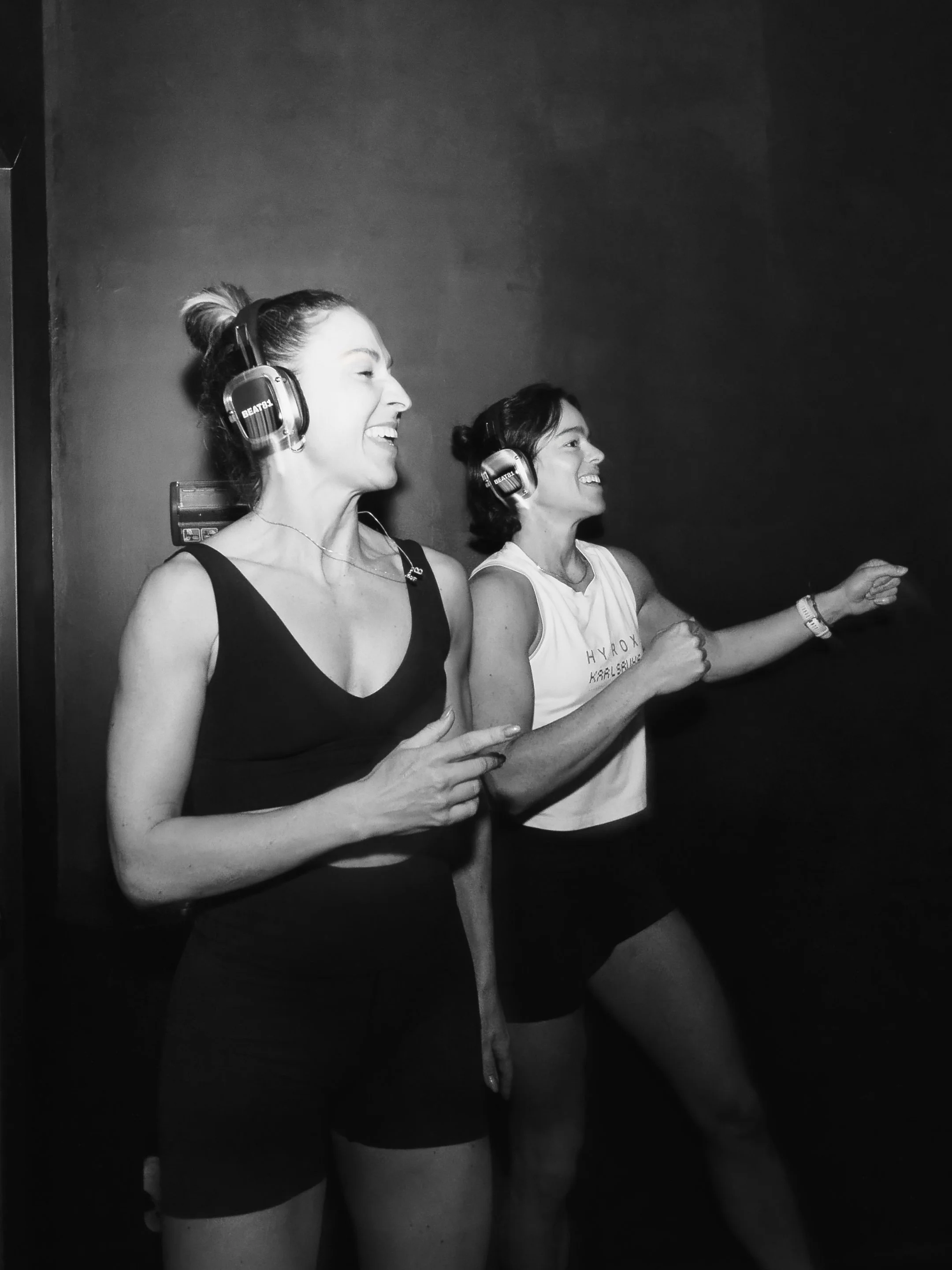 Zwei Frauen tragen Kopfhörer und Tänzer-Tops, während sie tanzen und lachen in einer dunklen Umgebung