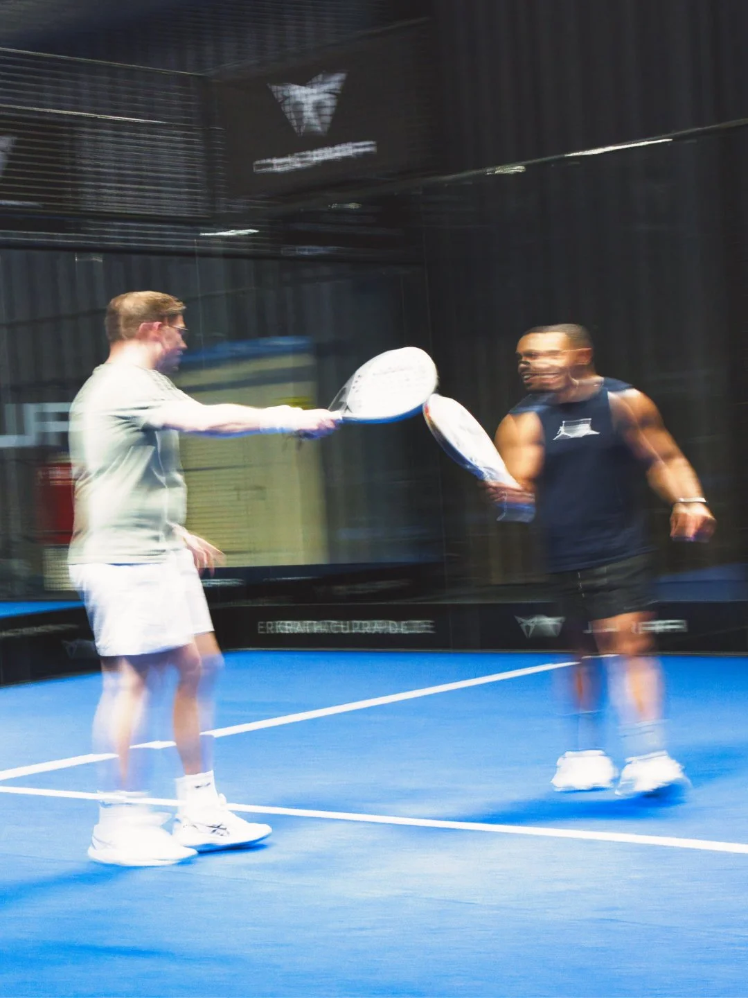 Zwei Männer spielen Padel Indoor, eine Person in Weiß, die andere in Dunkelblau, scheinen im Spiel zu sein