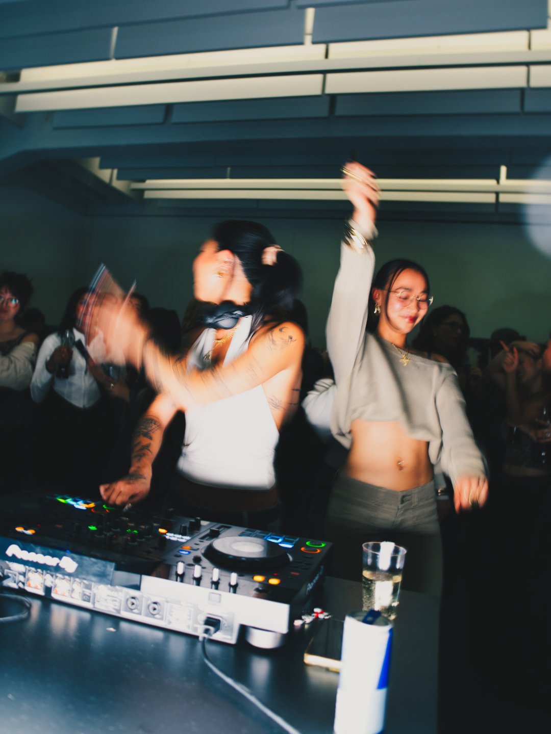 Zwei Frauen tanzen auf einer Party, vor einem DJ-Pult mit Musikgeräten, im Hintergrund eine Gruppe von Menschen, eine Frau hält ein Glas Wasser, die Szene wirkt lebendig und energiegeladen
