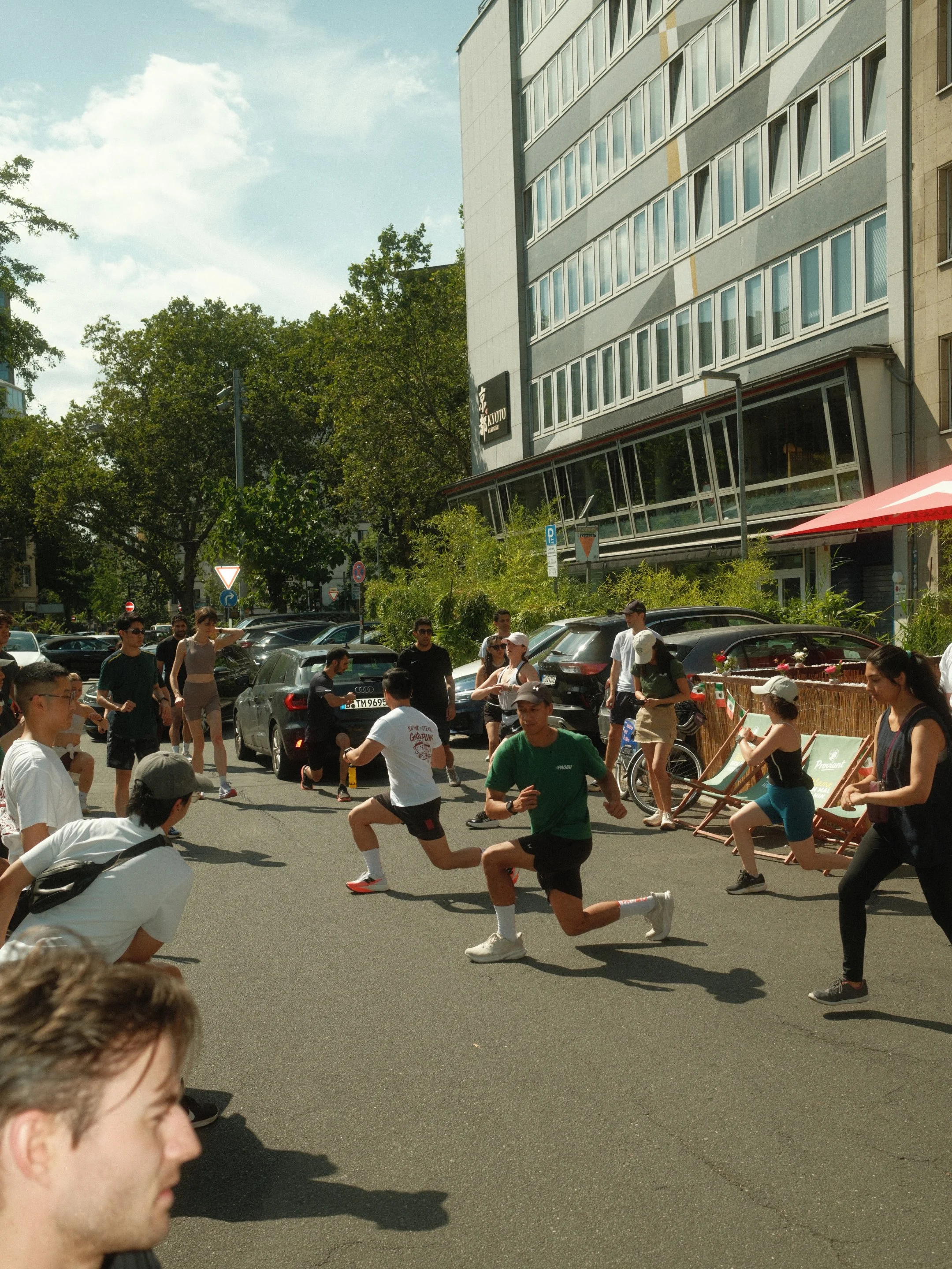 Menschen beim Laufen in einer Stadtstraße mit Autos, Gebäuden und Bäumen im Hintergrund, bei warmem Wetter