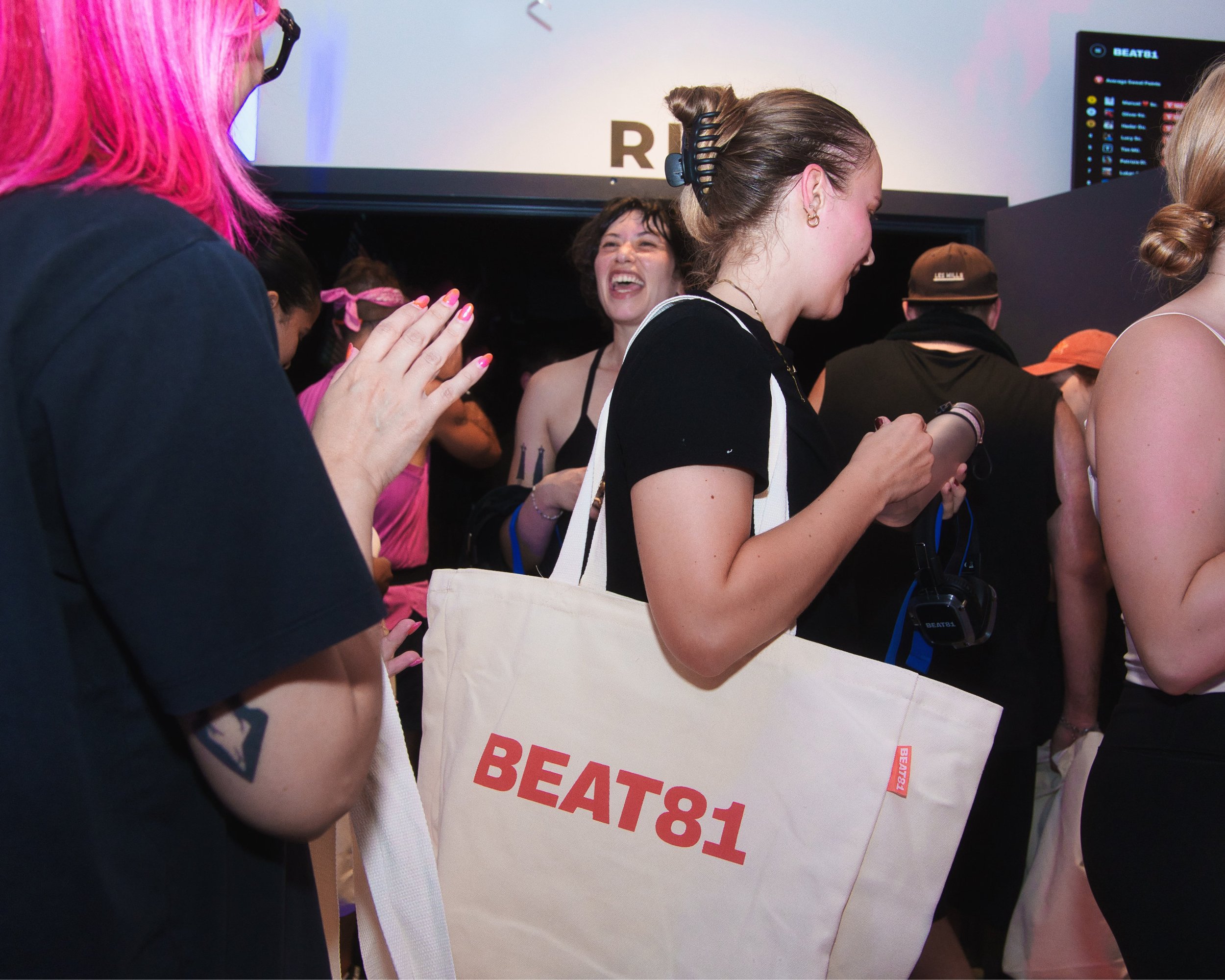 Mehrere Menschen bei einer Veranstaltung, einige tragen hörgeräte, einer hält ein Schild mit der Aufschrift 'BEATS81', im Hintergrund ein Bildschirm