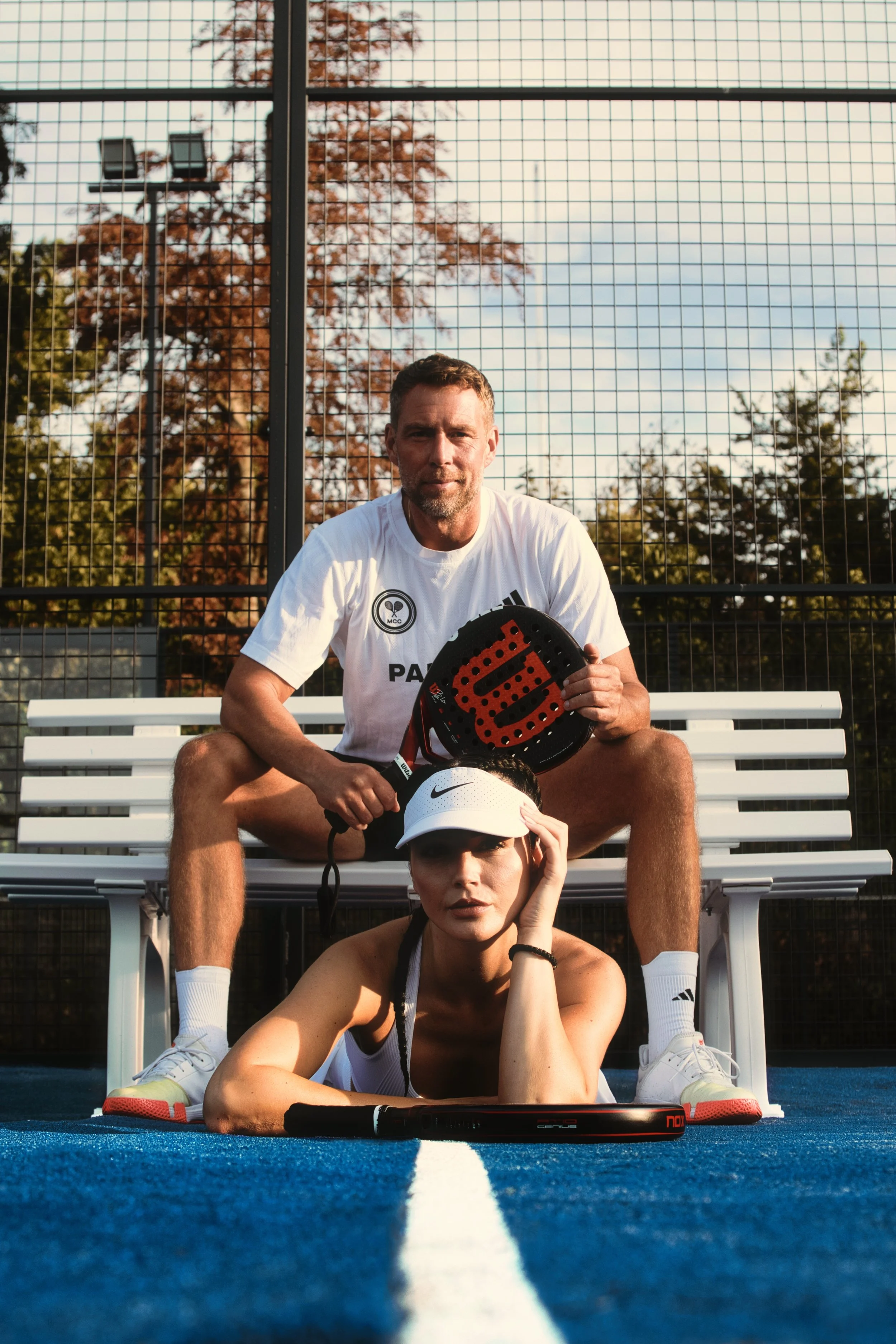 Zwei Menschen auf einem Padelplatz, einer sitzt auf einer Bank und hält einen Schläger, eine Person liegt auf dem Boden und trägt eine weiße Cap, die andere sitzt auf der Bank mit einem Tennisball in der Hand