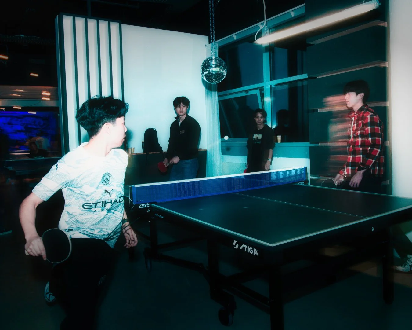 Zwei junge Männer spielen Tischtennis in einem modernen Raum, während zwei andere Personen im Hintergrund zuschauen