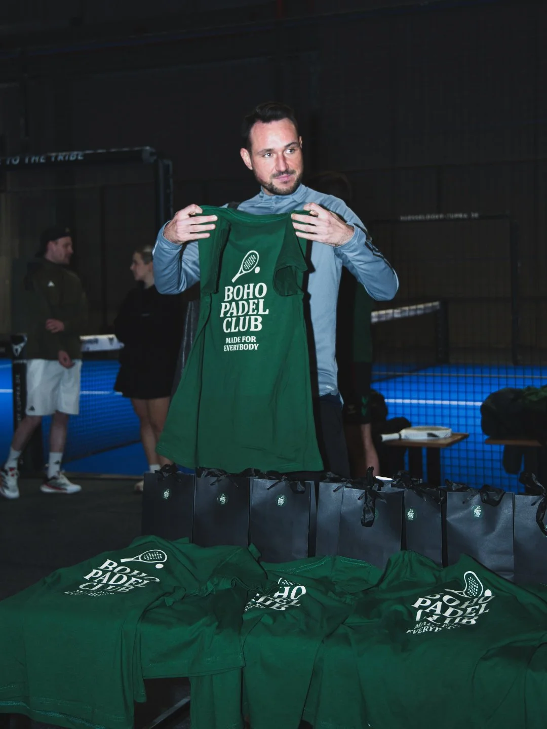 Ein Mann hält ein grünes Paddel-Club-T-Shirt mit der Aufschrift 'BOHO PADEL CLUB MADE FOR EVERYBODY' vor seiner Brust in einem Indoor-Padelfeld. Auf dem Tisch und im Vordergrund liegen weitere grüne T-Shirts und schwarze Tragetaschen