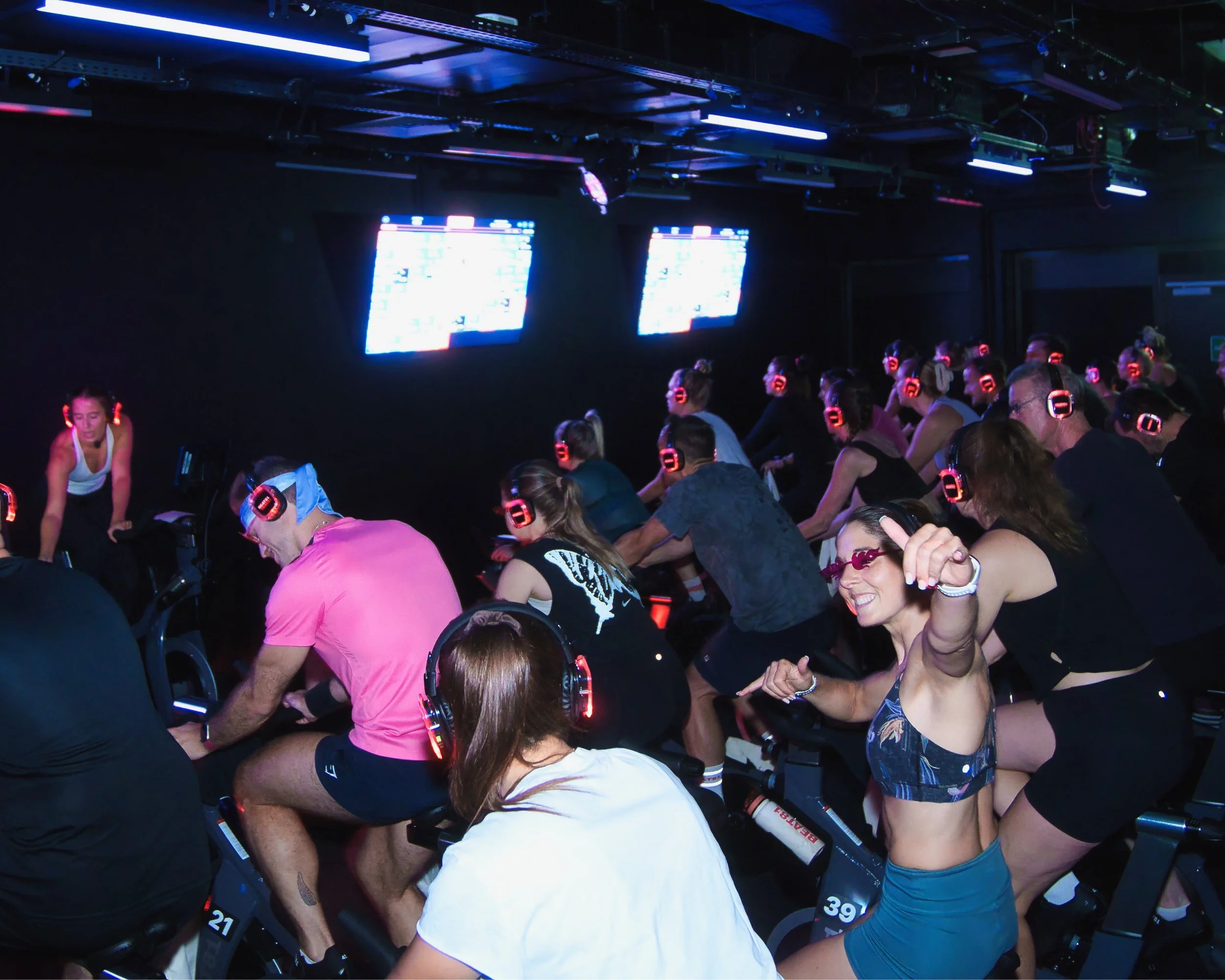 Menschen nehmen an einer festen Indoor-Spin-Klasse mit Headsets teil, während sie auf Spin-Bikes trainieren, in einem dunklen Raum mit digitalen Bildschirmen und blauer Beleuchtung
