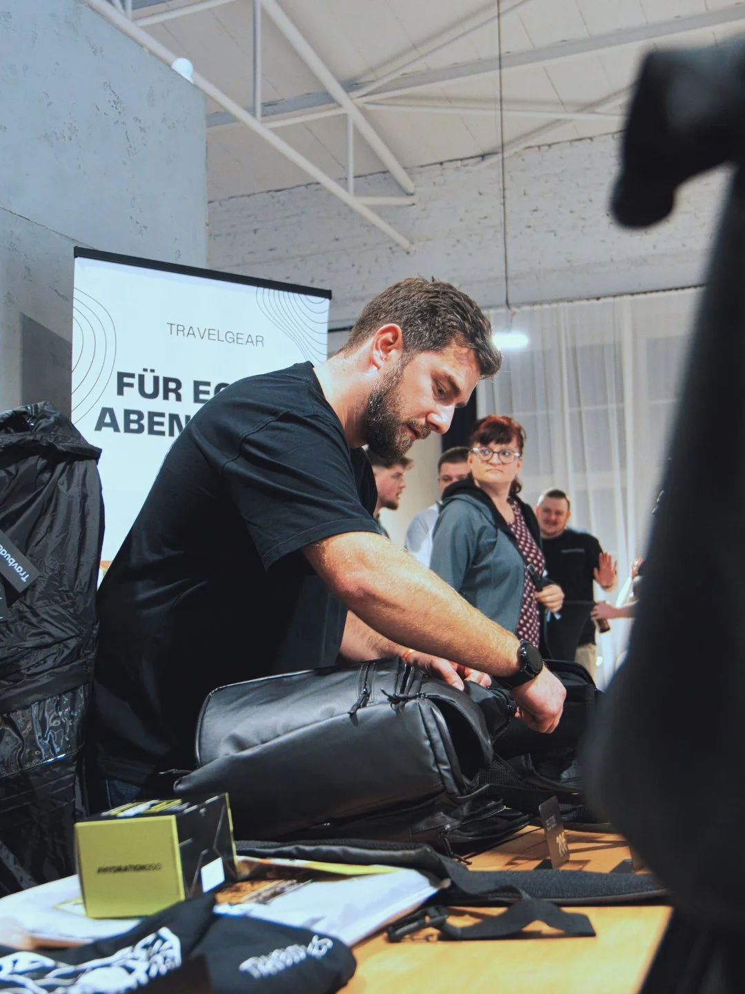 Mann packt eine schwarze Tasche auf einem Tisch, hinter ihm stehen mehrere Menschen in einem Raum mit weißen Wänden und einer Projektionsleinwand
