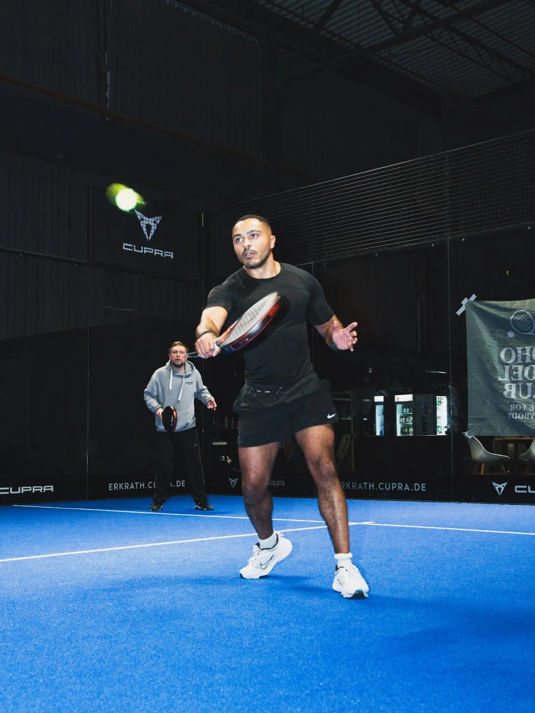 Zwei Männer spielen Padel Indoor. Der Mann im Vordergrund trägt schwarze Kleidung und weiße Schuhe, er hält einen Tennisschläger in der Hand