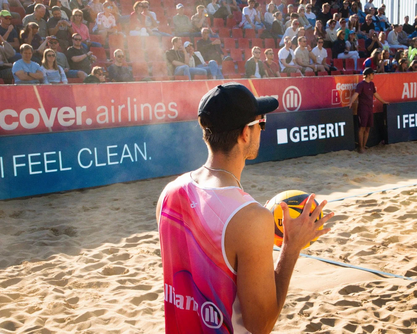 Ein männlicher Beachvolleyballspieler bereitet sich zum Aufschlag vor, trägt eine pinkfarbene Weste, eine schwarze Mütze und Sonnenbrille, während das Publikum im Hintergrund das Spiel beobachtet