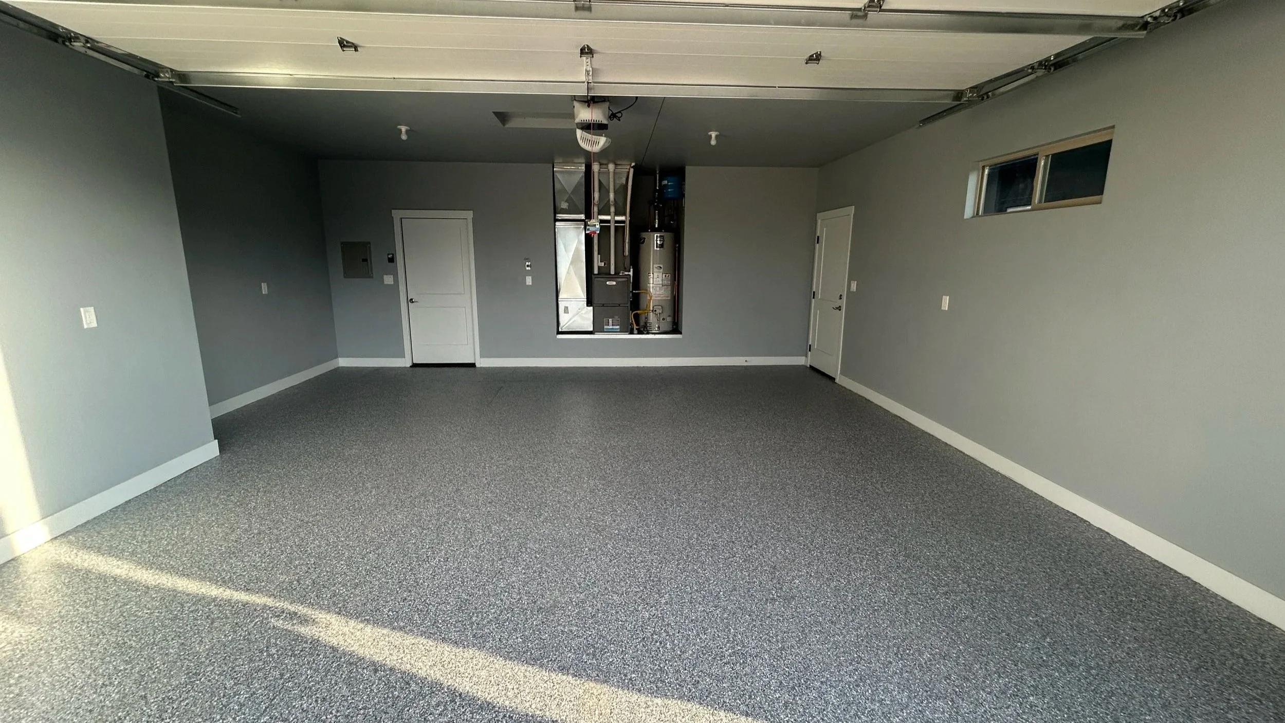 Empty garage with gray walls, gray carpeted floor, and a white ceiling with a garage door opener mechanism. There are two closed doors, a small horizontal window, and an open panel revealing HVAC equipment.