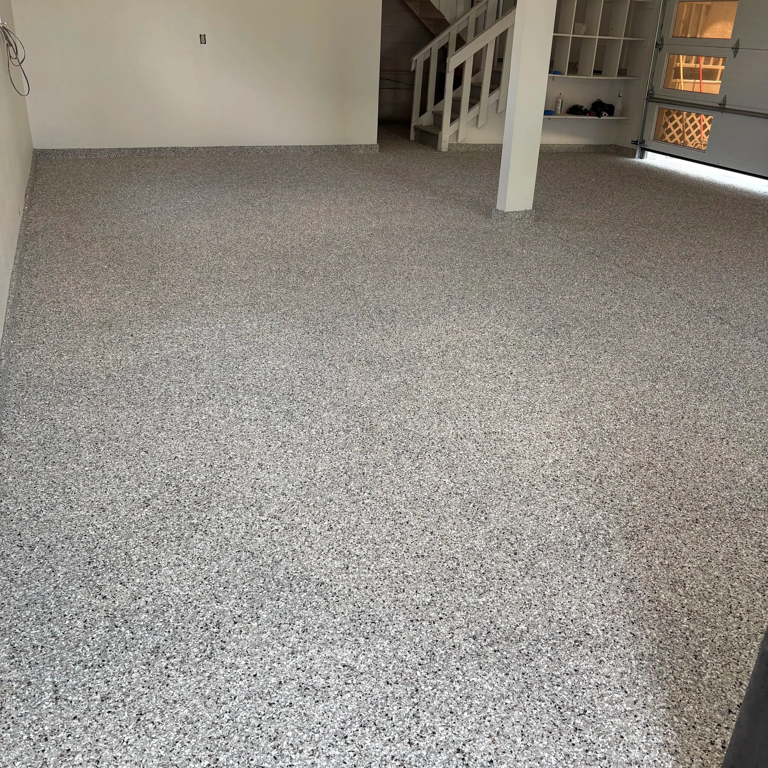 Empty garage with speckled epoxy-coated concrete floor, white walls, a staircase, and a partially open garage door revealing a construction area outside.